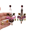 Rosey gold Festive Drop statement earrings shown hanging against a white background with my hand behind for scale.