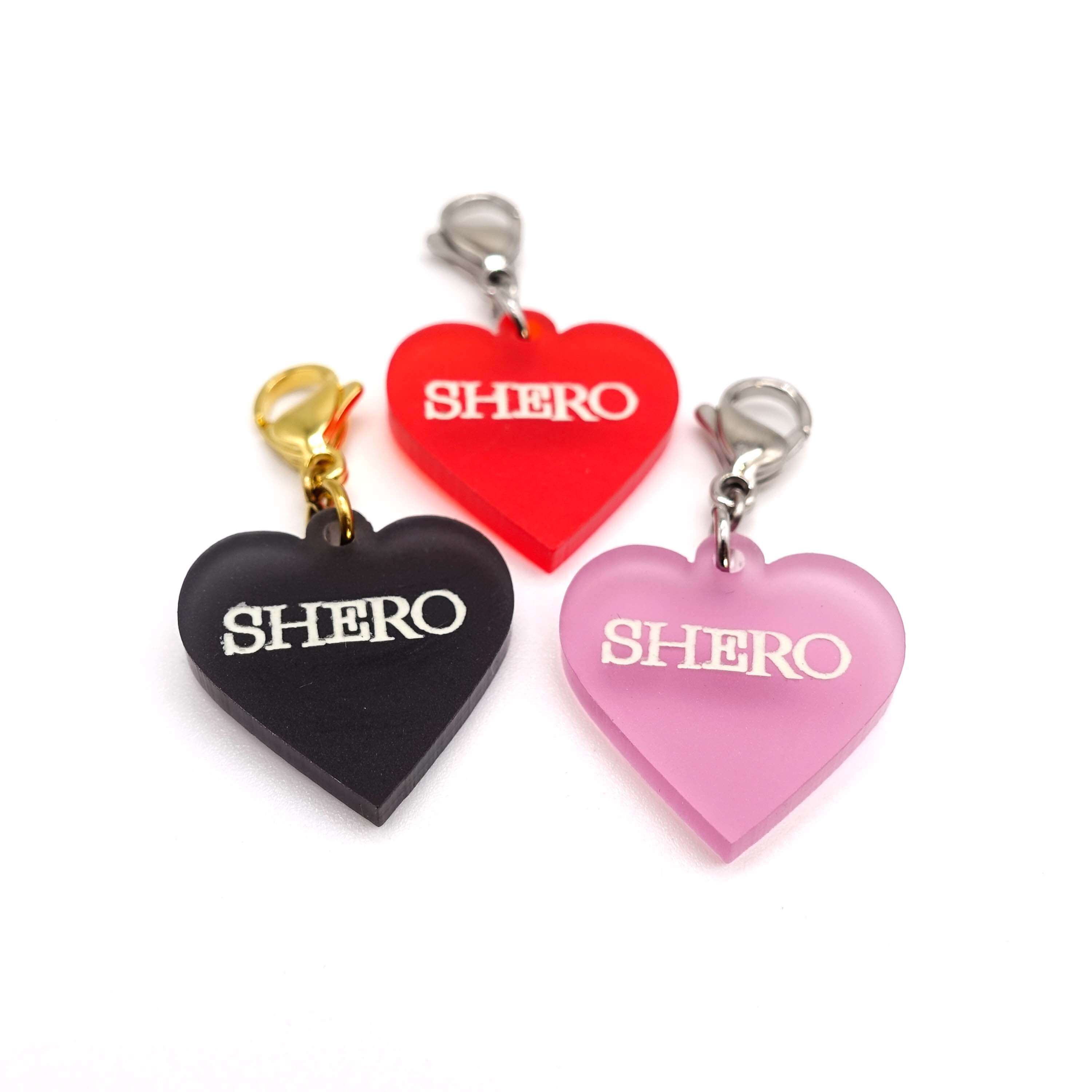 Heart charms in chilli frost, pink frost and slate frost etched in glow-in-the-dark pigment with the word 'Shero'