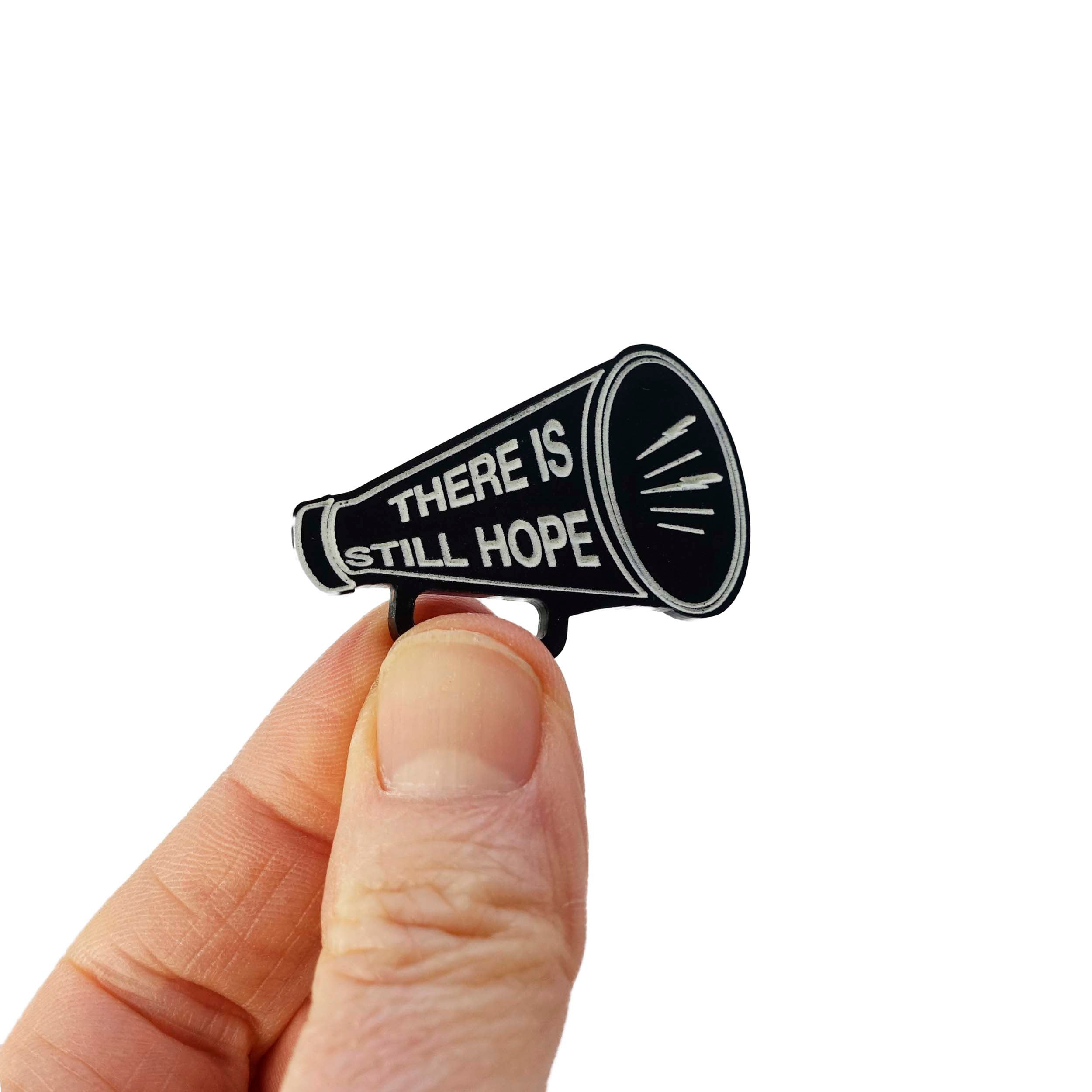 Matte black small megaphone brooch etched in glow-in-the-dark pigment with the words There is Still Hope. Shown held up in my fingers for scale.