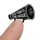Small megaphone brooch etched with the words Take them to the Hague in glow-in-the-dark-pigment, shown held up in my fingers against a white background.