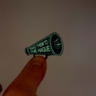 Small megaphone brooch etched with the words Take them to the Hague in glow-in-the-dark-pigment, shown held up in my fingers glowing in the dark.