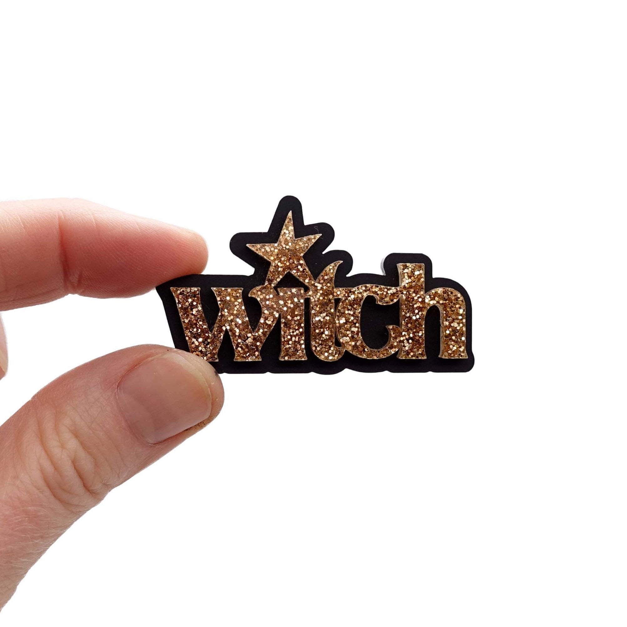 Witch brooch in gold glitter, shown held up in Sarah's fingers for scale. £2 goes to Welsh Women's Aid.
