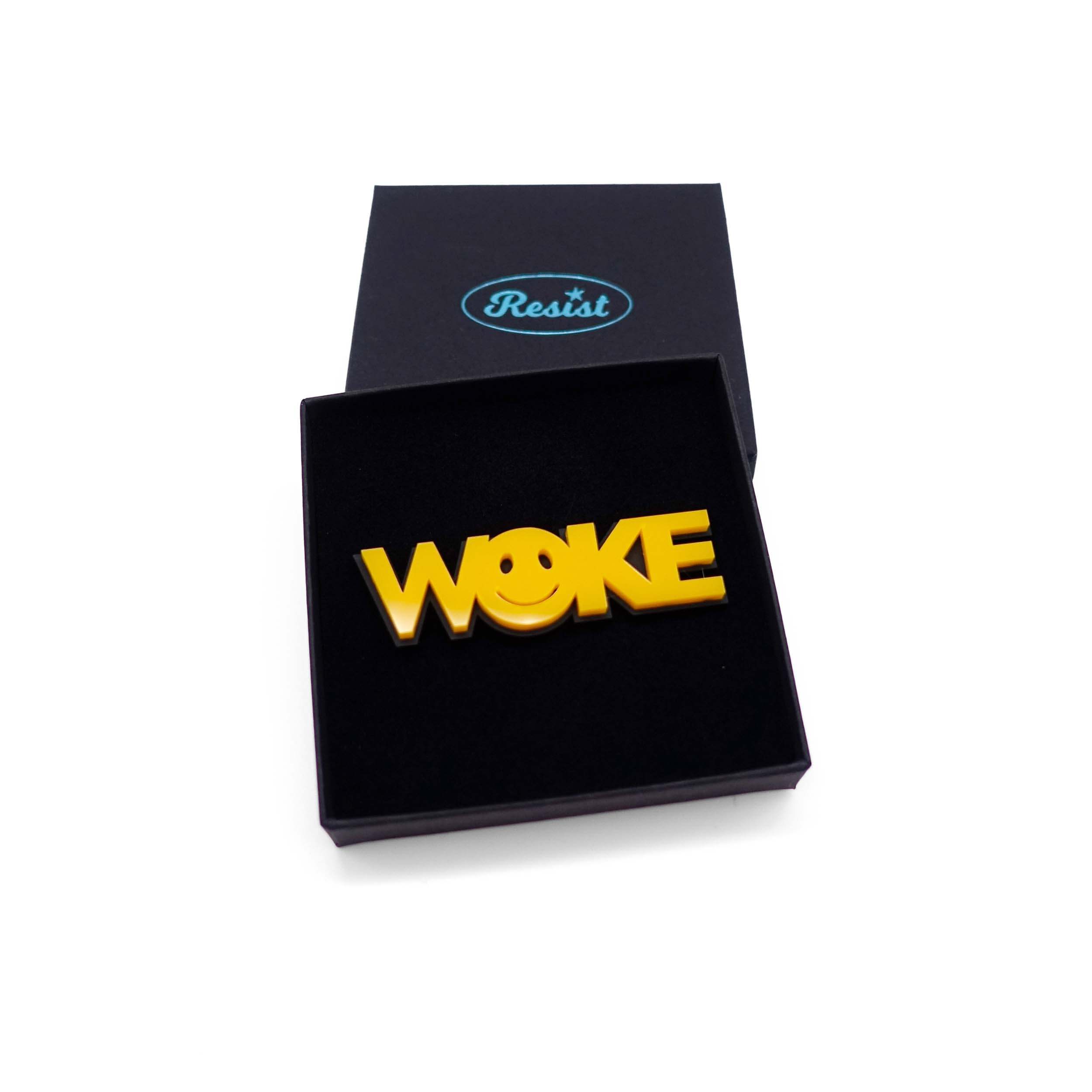 A yellow WOKE brooch with a 90s smiley face in the O, shown in a Wear and Resist gift box.