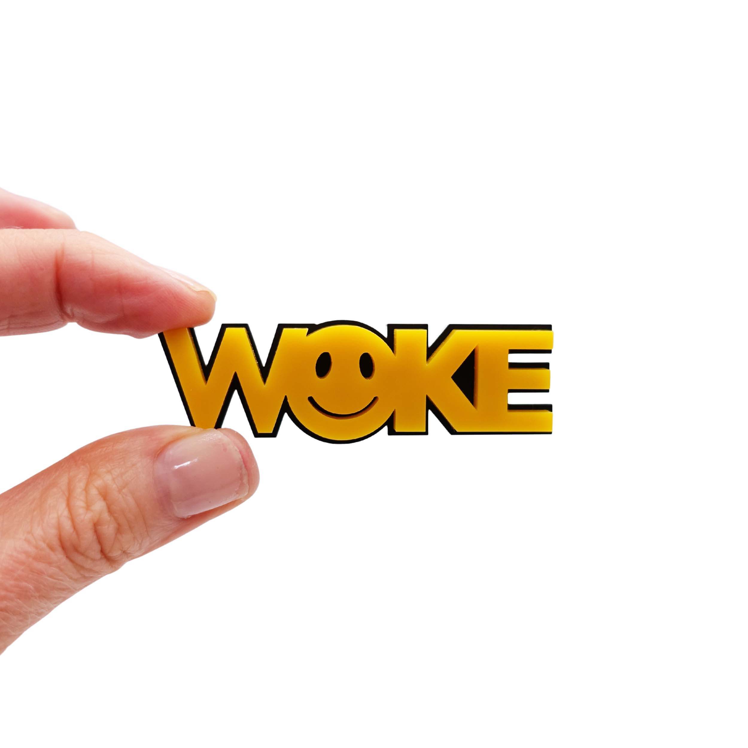 My hand holding a yellow brooch with the word 'WOKE' in bold letters and a 90s smiley face in the O.