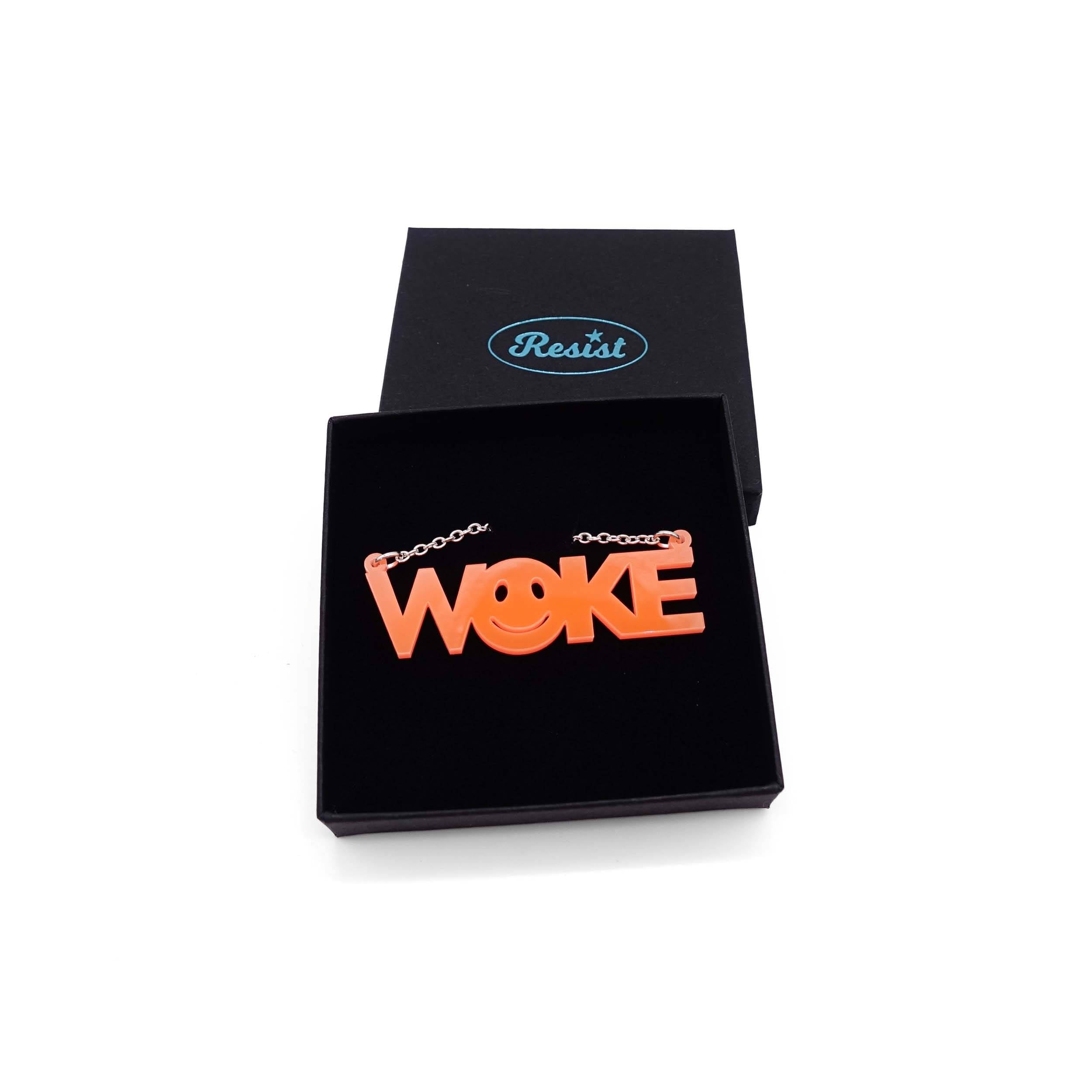 A flouro orange WOKE necklace with a 90s smiley face cut out of the O shown in a Wear and Resist gift box.
