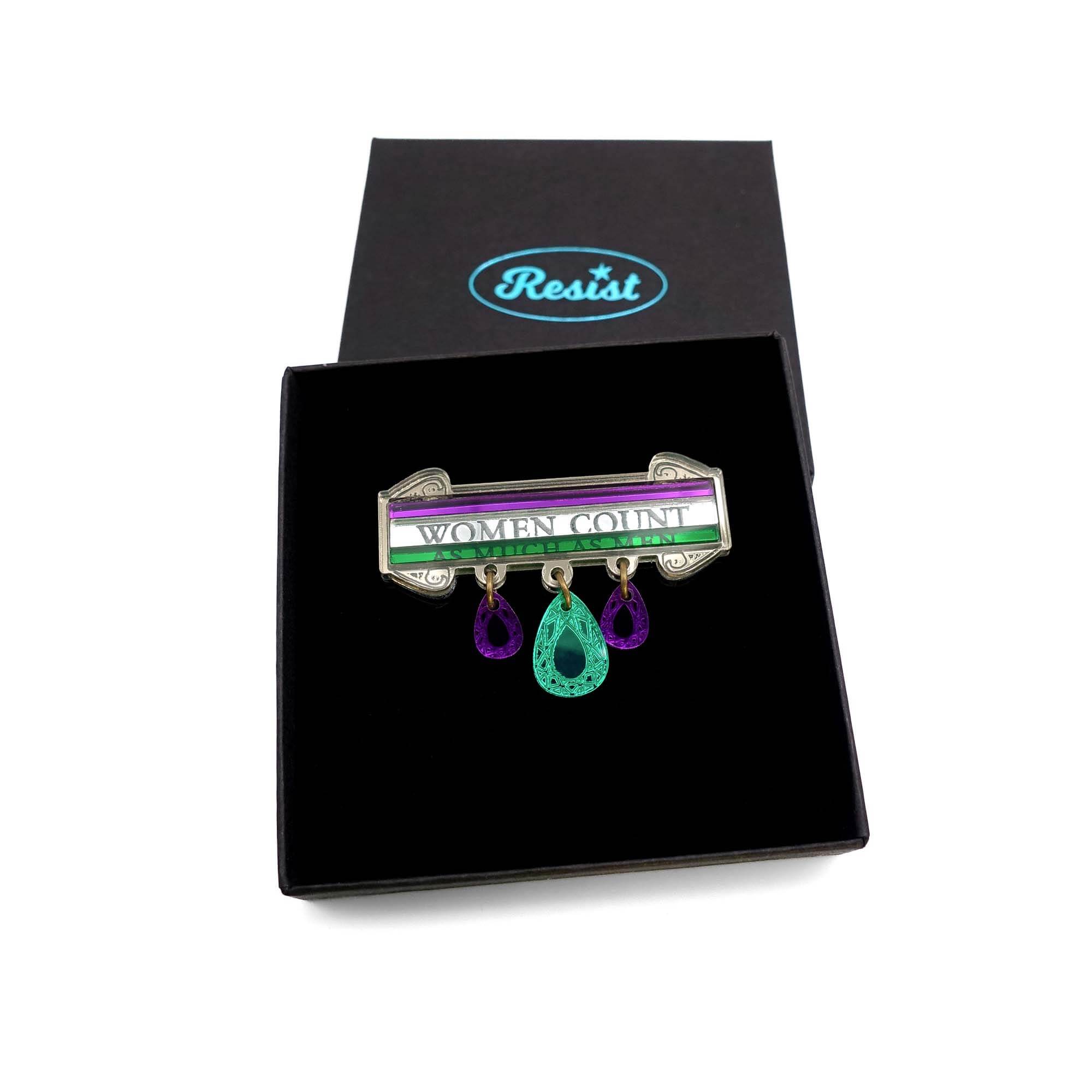 A brooch made in acrylic to look like etched bronze with the words Women Count and As Much as Men etched on the three bands in the Suffragette colours, with jewels hanging down in green and purple.