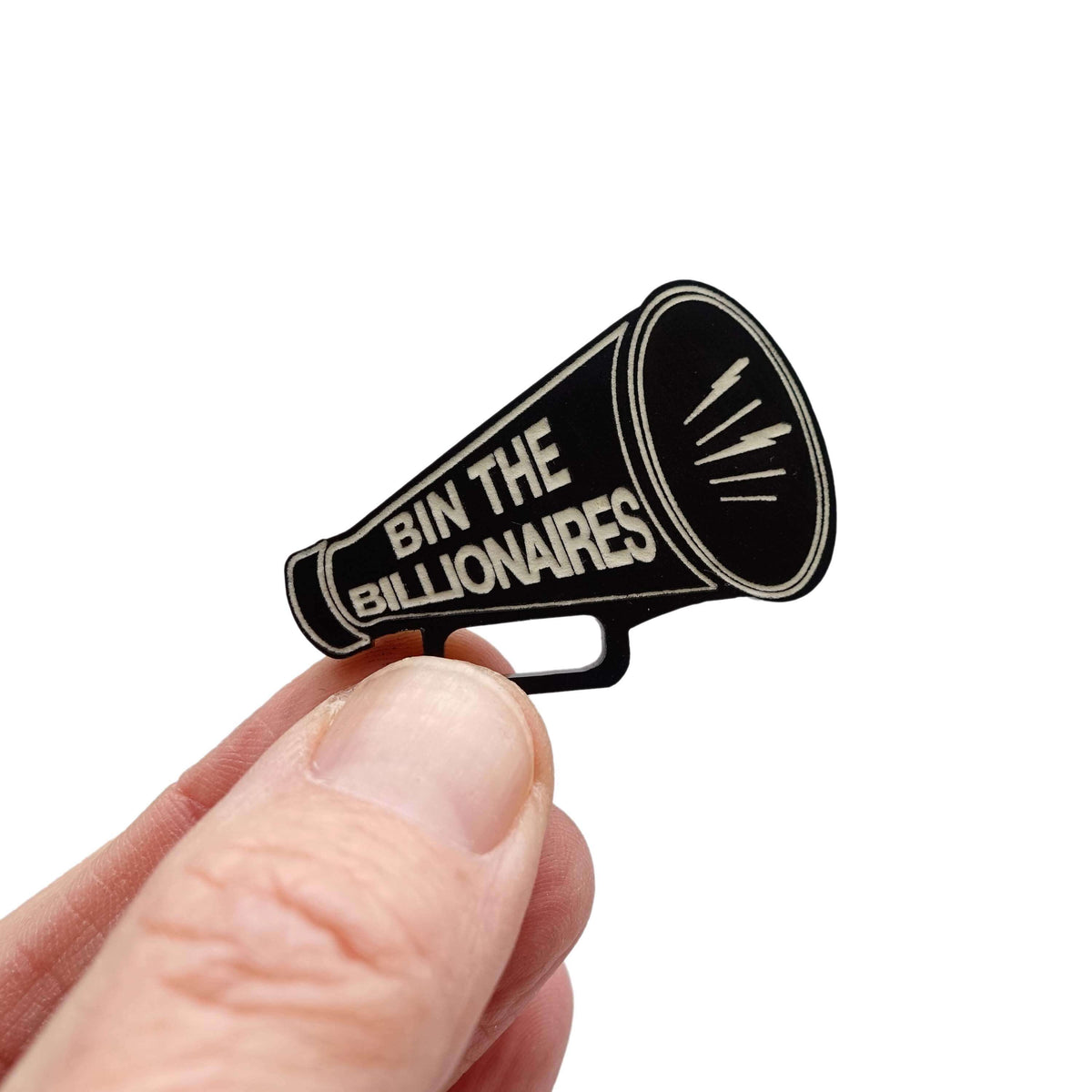 A matte black acrylic brooch in the shape of a megaphone, etched with glow in the dark paint with the words BIN THE BILLIONAIRES, shown held up in my fingers.