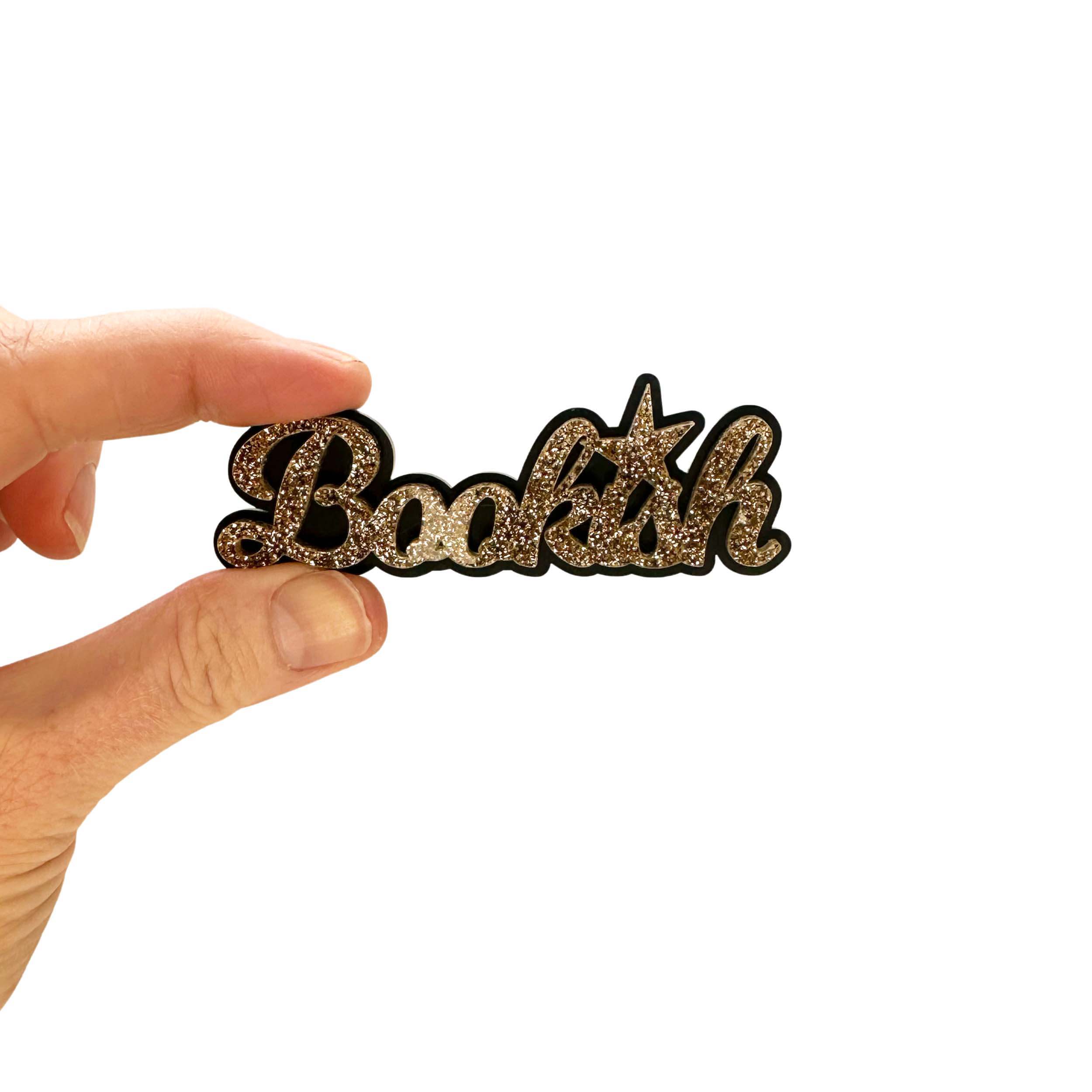 Bookish brooch in gold glitter shown held up against a white background.