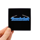 Matte bright blue Feminist necklace shown held up in a Wear and Resist gift box.