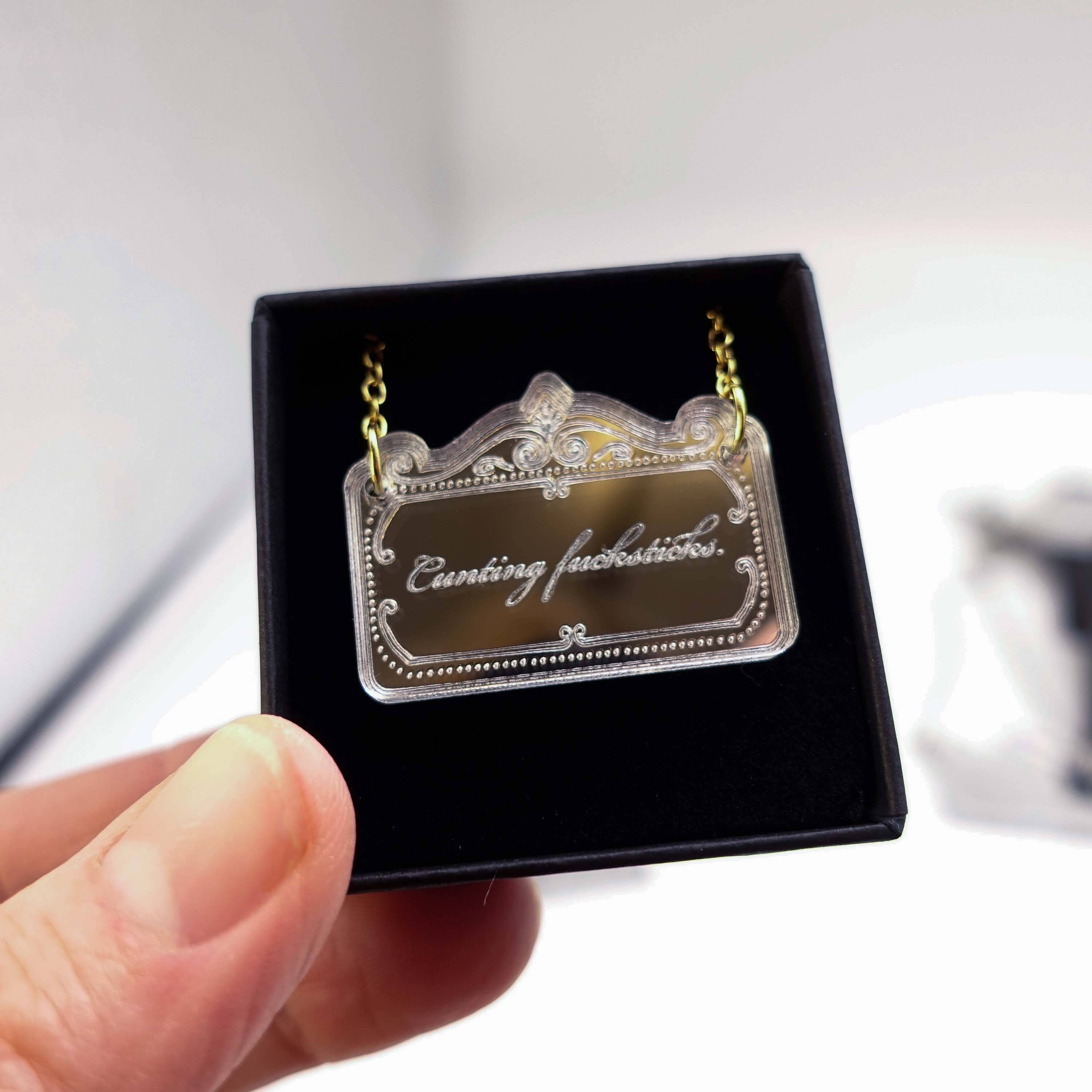 A delicate necklace etched with 'c*nting fucksticks' shown held up in a small Wear & Resist gift box.
