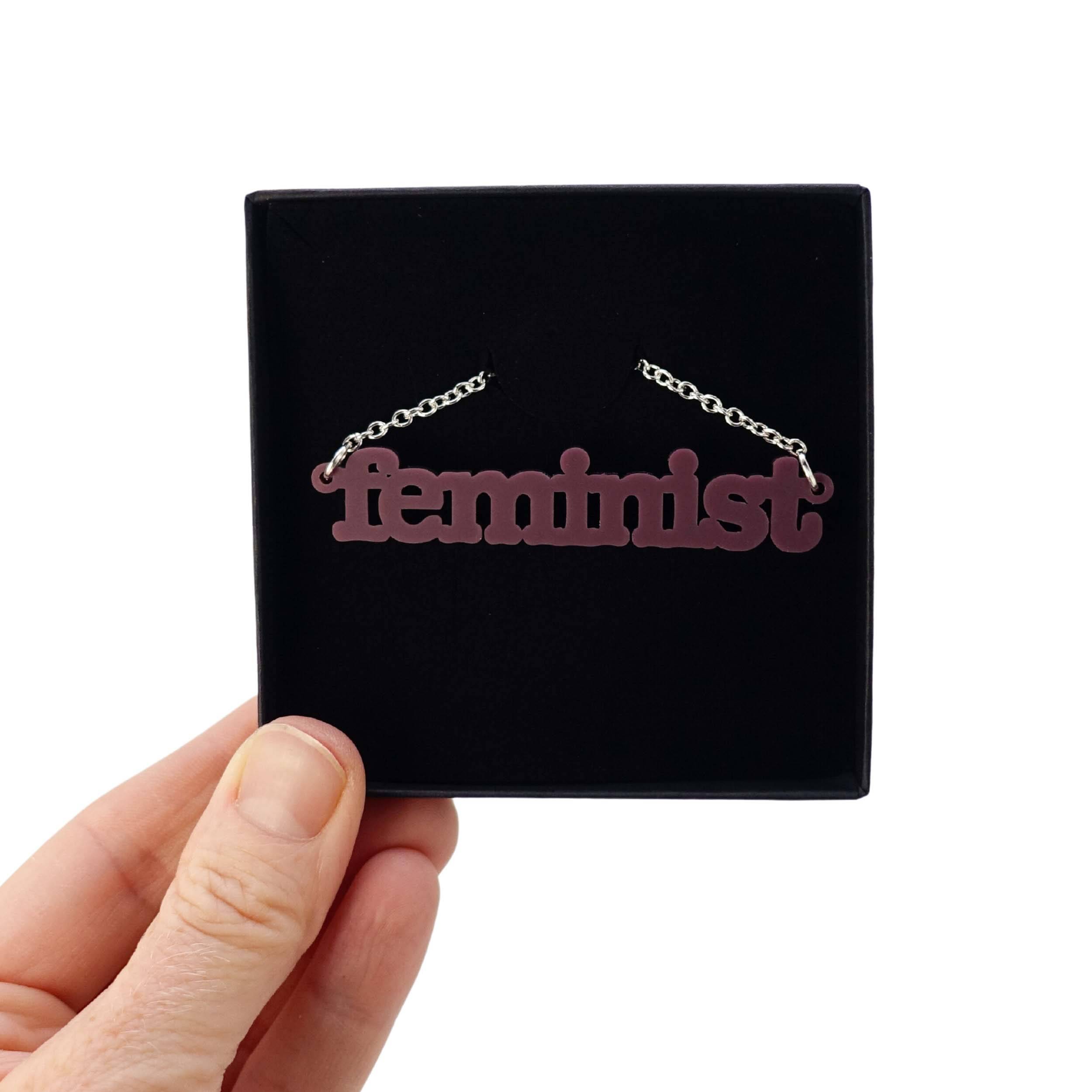 Cassis Feminist necklace shown held up in a Wear and Resist gift box.