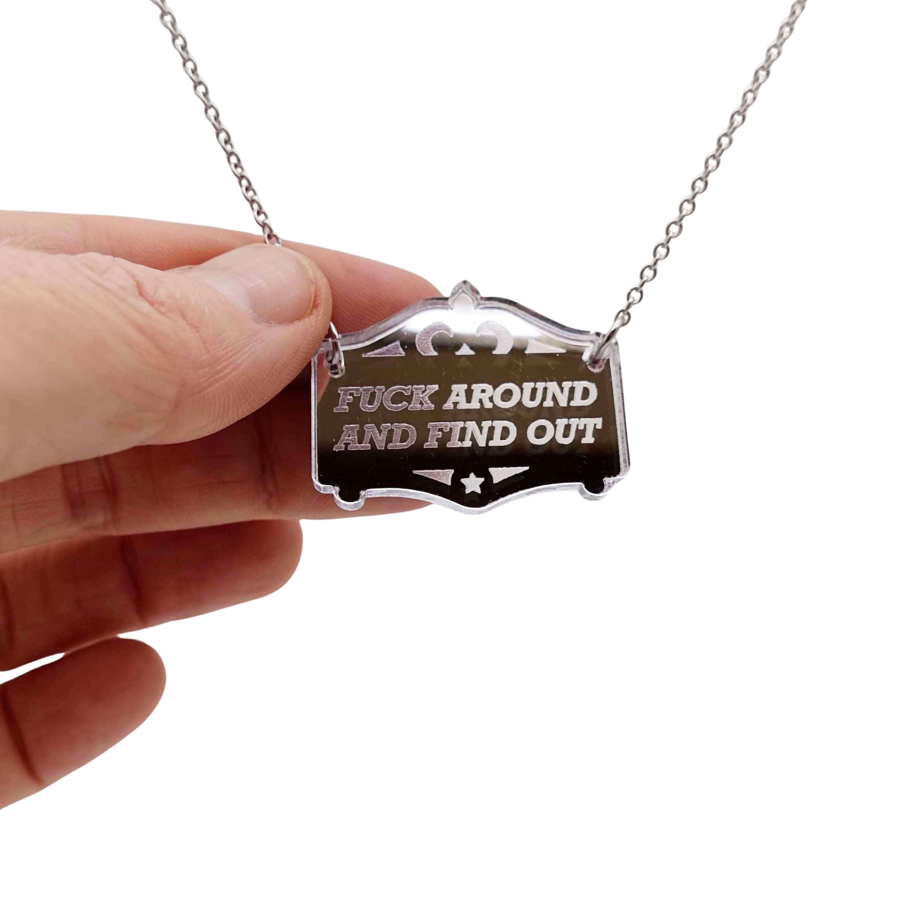 F*ck around and find out mirror cowboy necklace in silver, shown hanging on a stainless steel chain with my hand for scale.