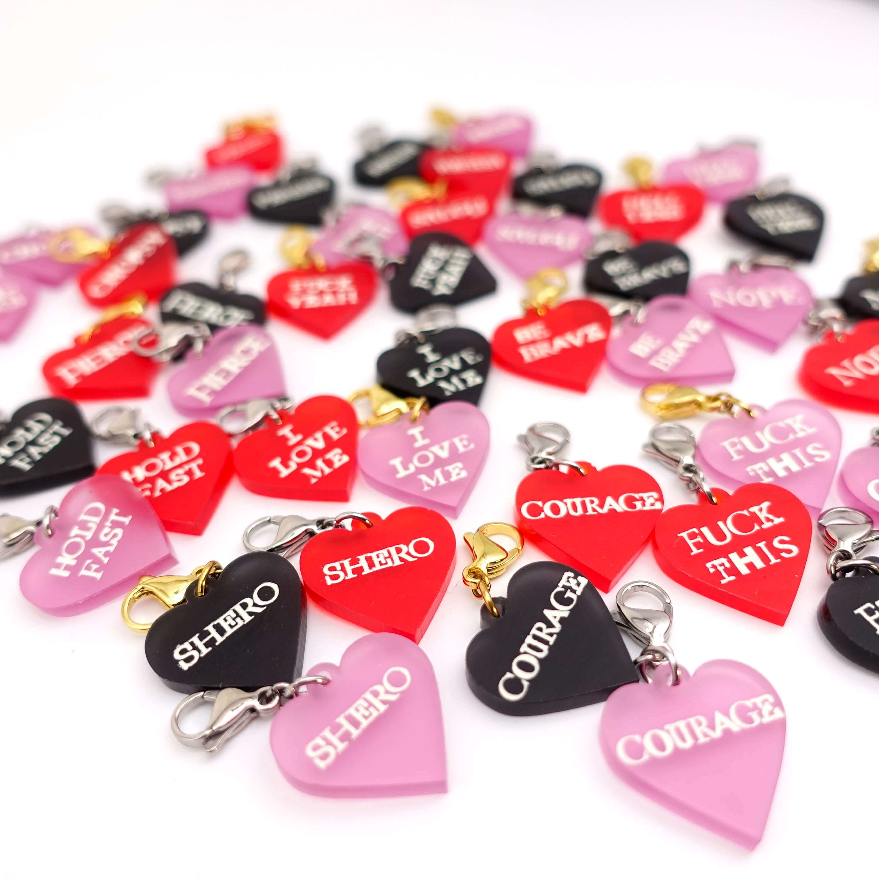 Heart shaped charmed in pink red and slate frost etched in glow-in-the-dark pigment with different words including: courage, shero, I love me, be brave, Fuck this, Fuck yeah, hold fast and nope.