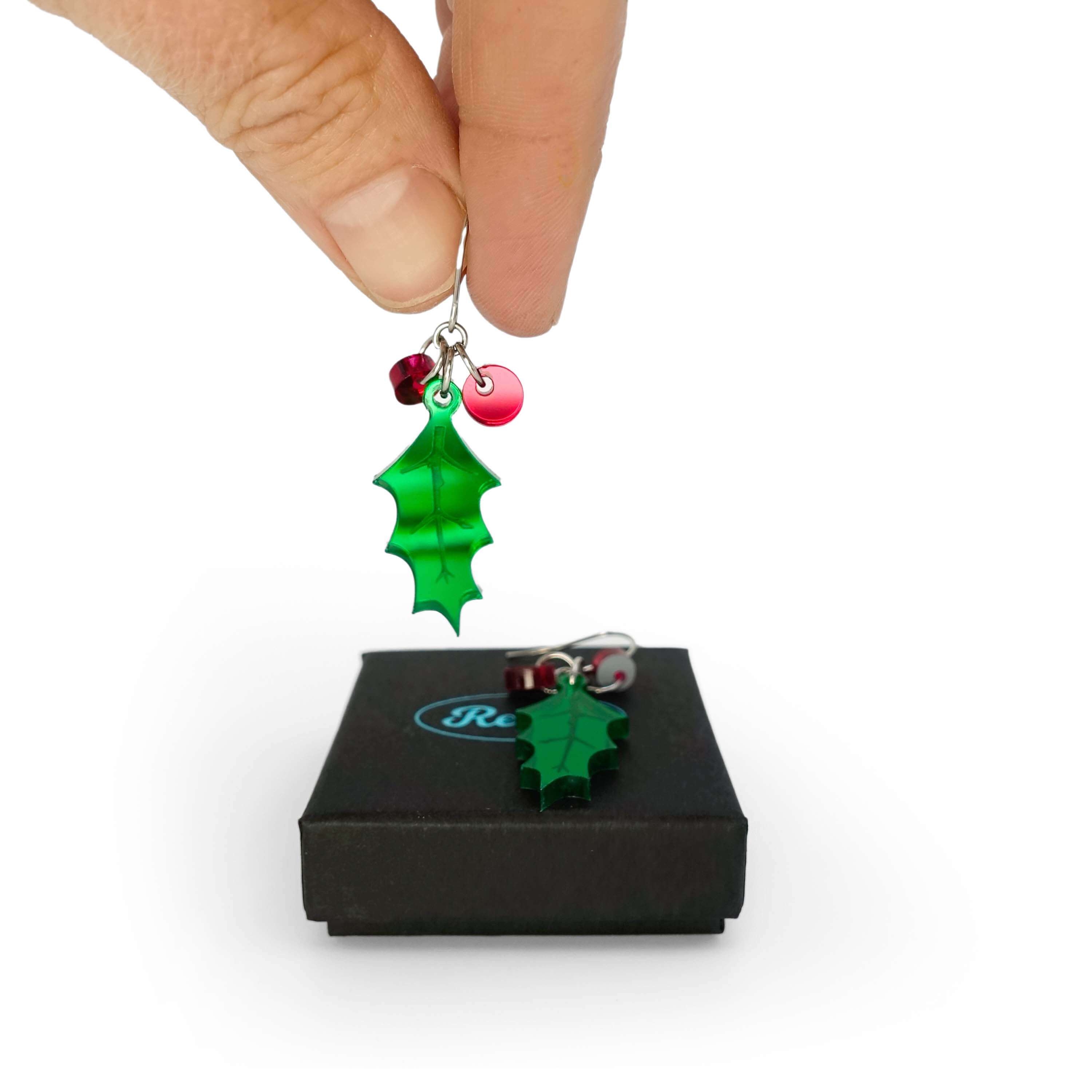 My hand holding one of a pair of naturalistic Holly earrings with red berries, the other one lying on a Wear and Resist gift box.