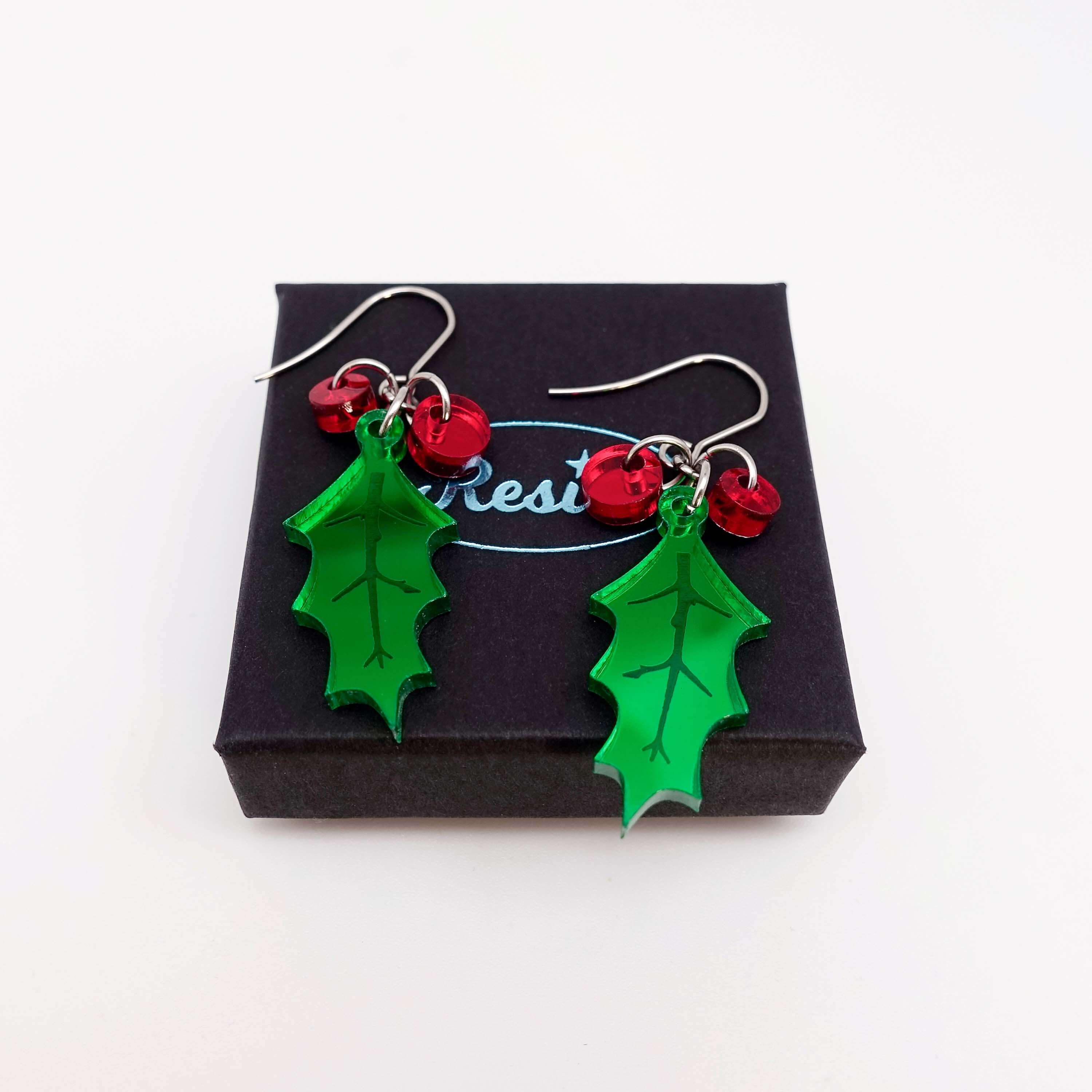 A pair of naturalistic Holly earrings with red berries shown on a small Wear and Resist gift box.