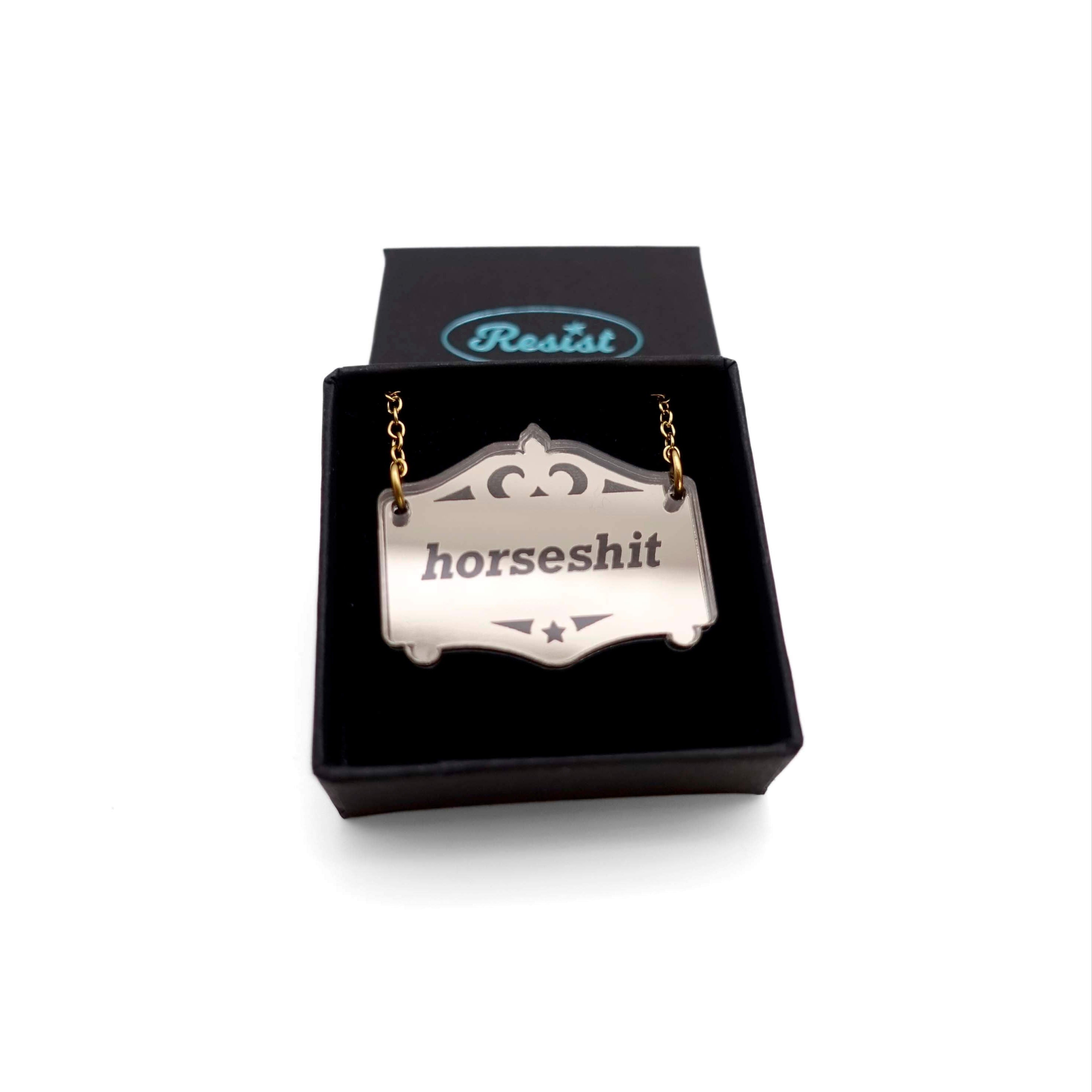 A bronze Mirror Cowboy Horsesh*t necklace, shown in a small Wear and Resist gift box.