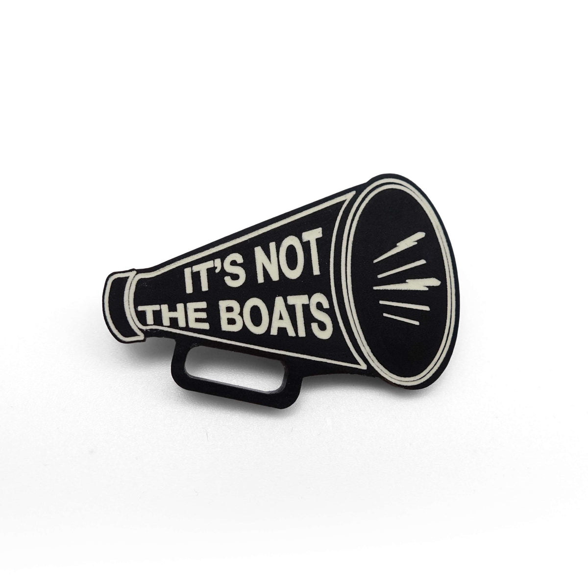 A matte black acrylic brooch in the shape of a megaphone etched in glow-in-the-dark pigment with the words 'It's not the boats', shown on a white background.