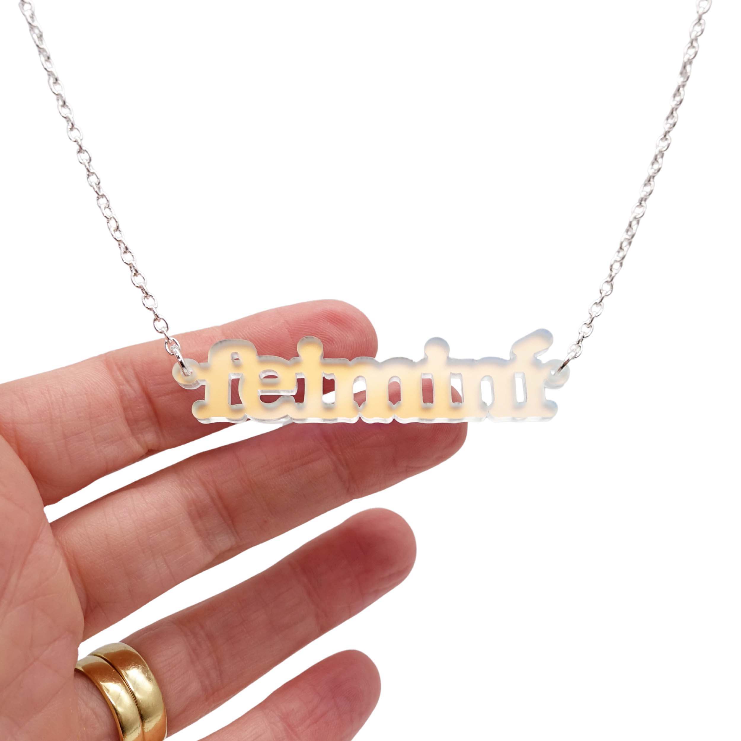 Irish Gaelic Feiminí necklace in matte iridescent, shown hanging against a white background, with my hand behind to show the luminosity of the colour.