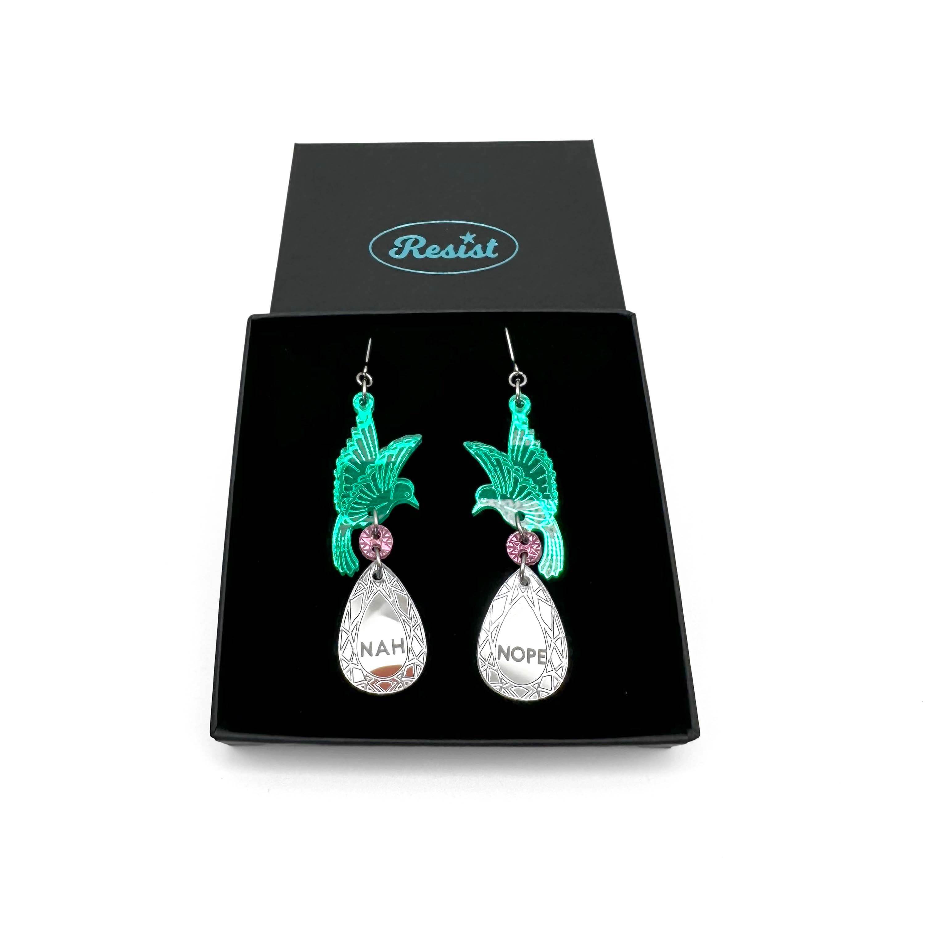 A pair of acrylic earrings with green birds holding silver 'jewels' that say 'Nah' and 'Nope'. Shown in a Wear & Resist gift box.
