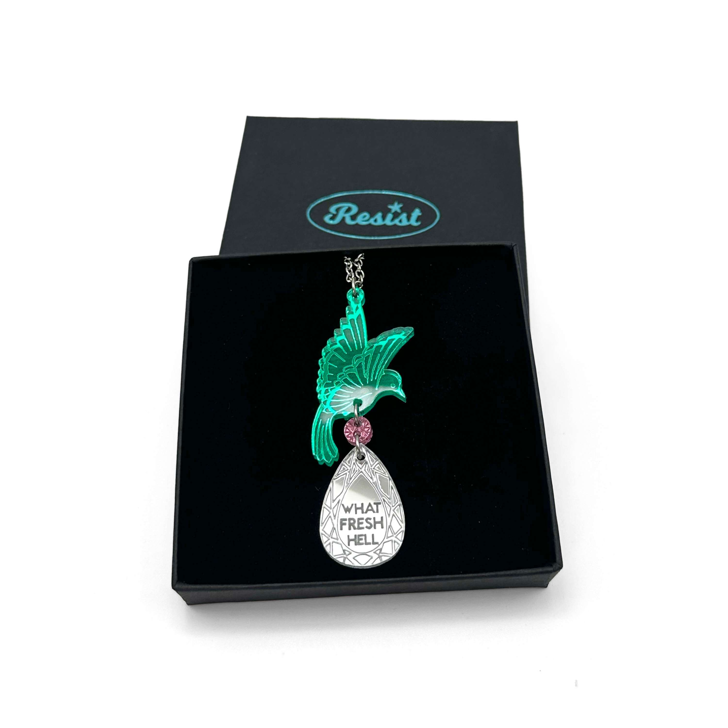An acrylic pendant necklace with a green bird holding a silver 'jewel' that says 'What Fresh Hell'. Shown in a Wear & Resist gift box.