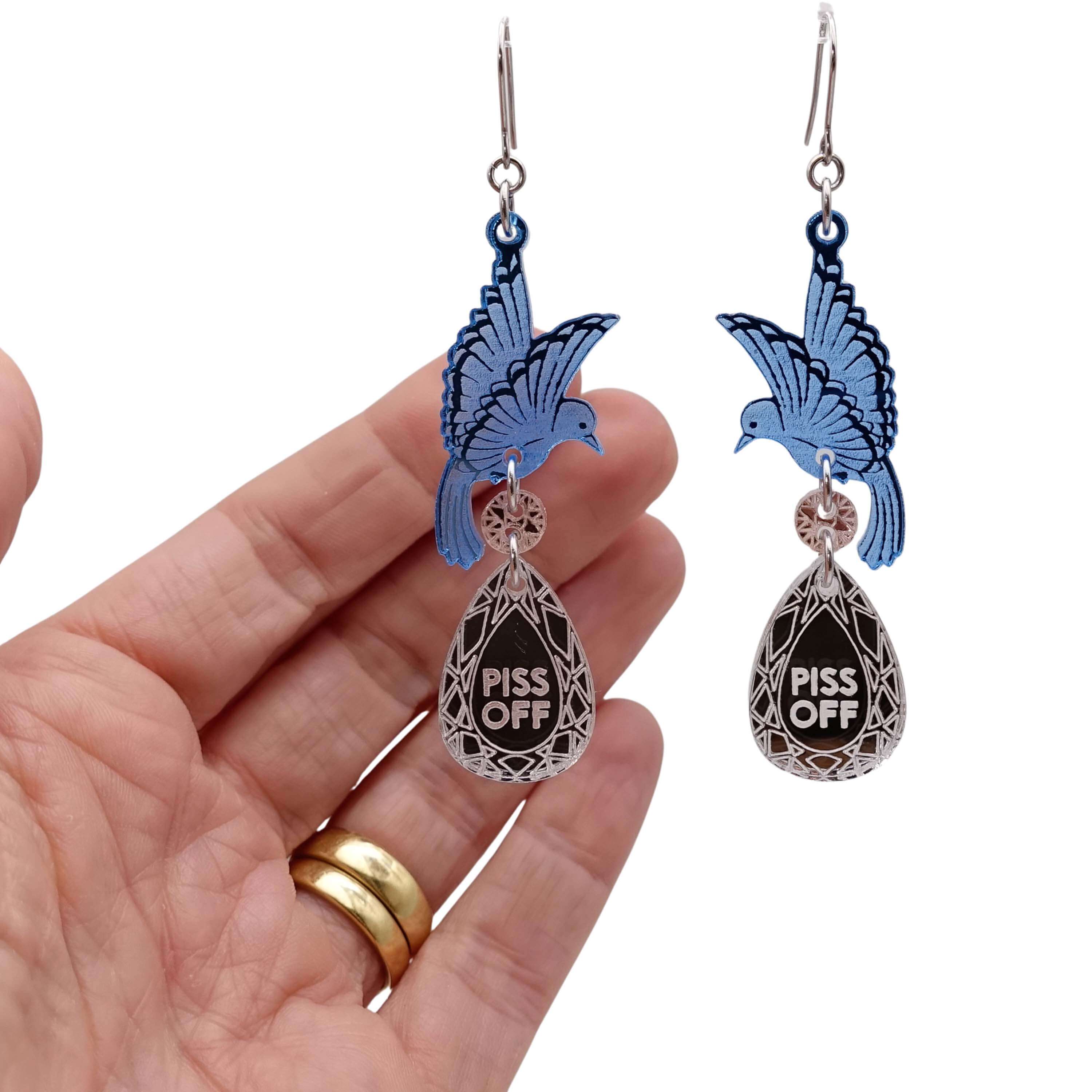 A pair of acrylic earrings with blue birds holding silver 'jewels' that say 'P*ss Off'. Photographed with my hand behind for scale.