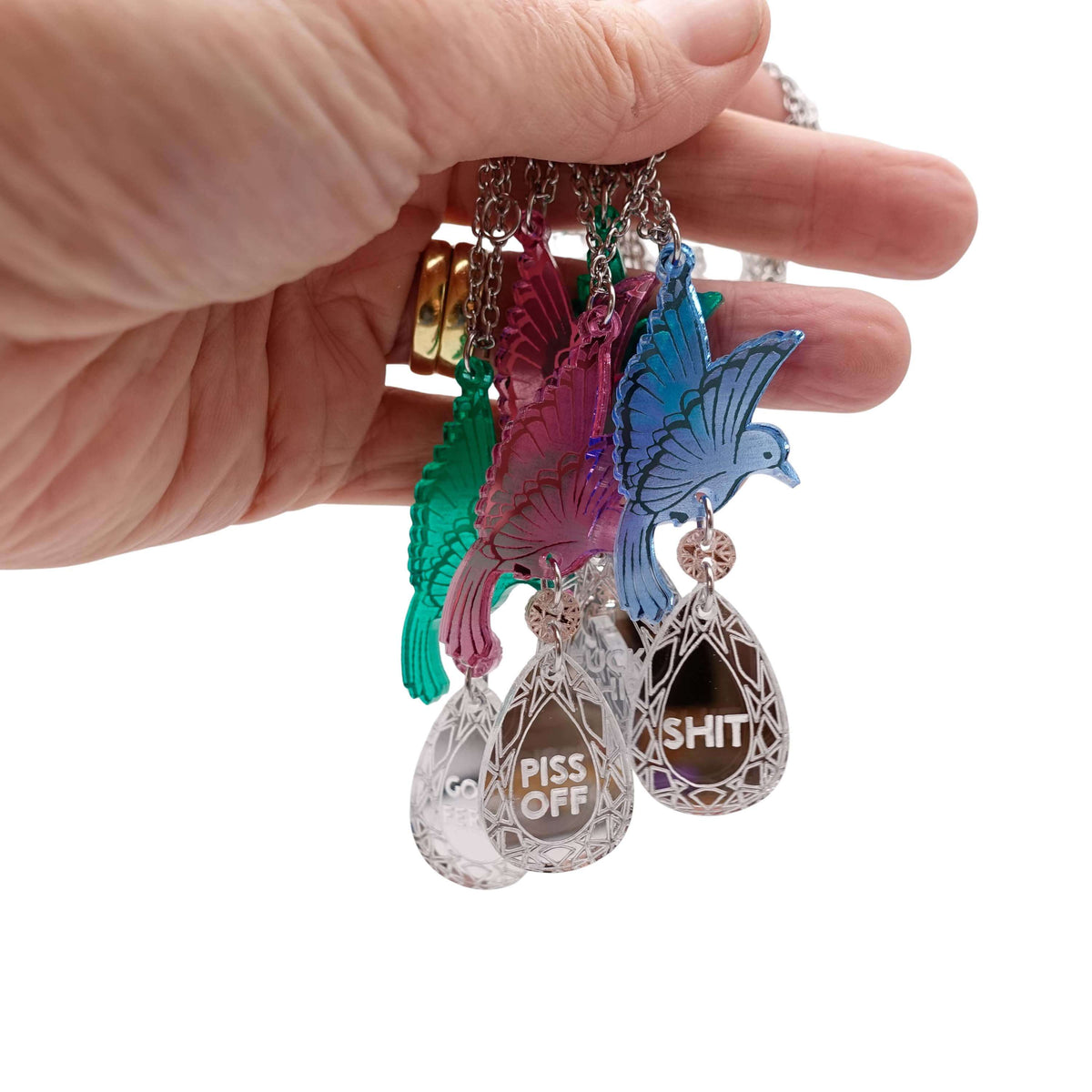 My hand holding a group of Rude Bird pendant necklaces in blue, pink and green, with sweary silver 'jewels'