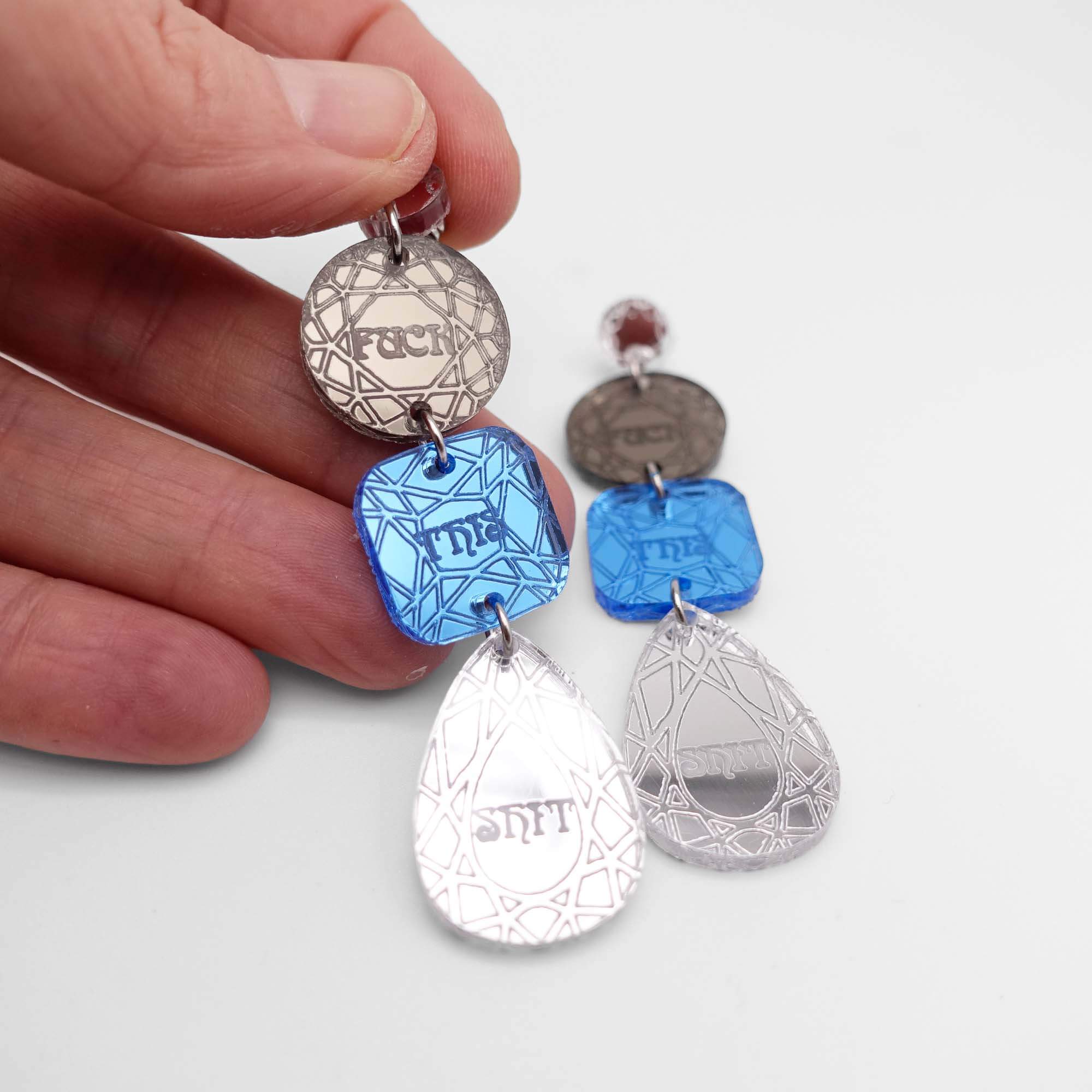 Sweary f*ck this sh*t earrings in bronze and sky, shown held up against a white background.