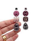 Sweary jewel drop earrings in pink, rose gold and silver, shown hanging against a white background with my hand behind.