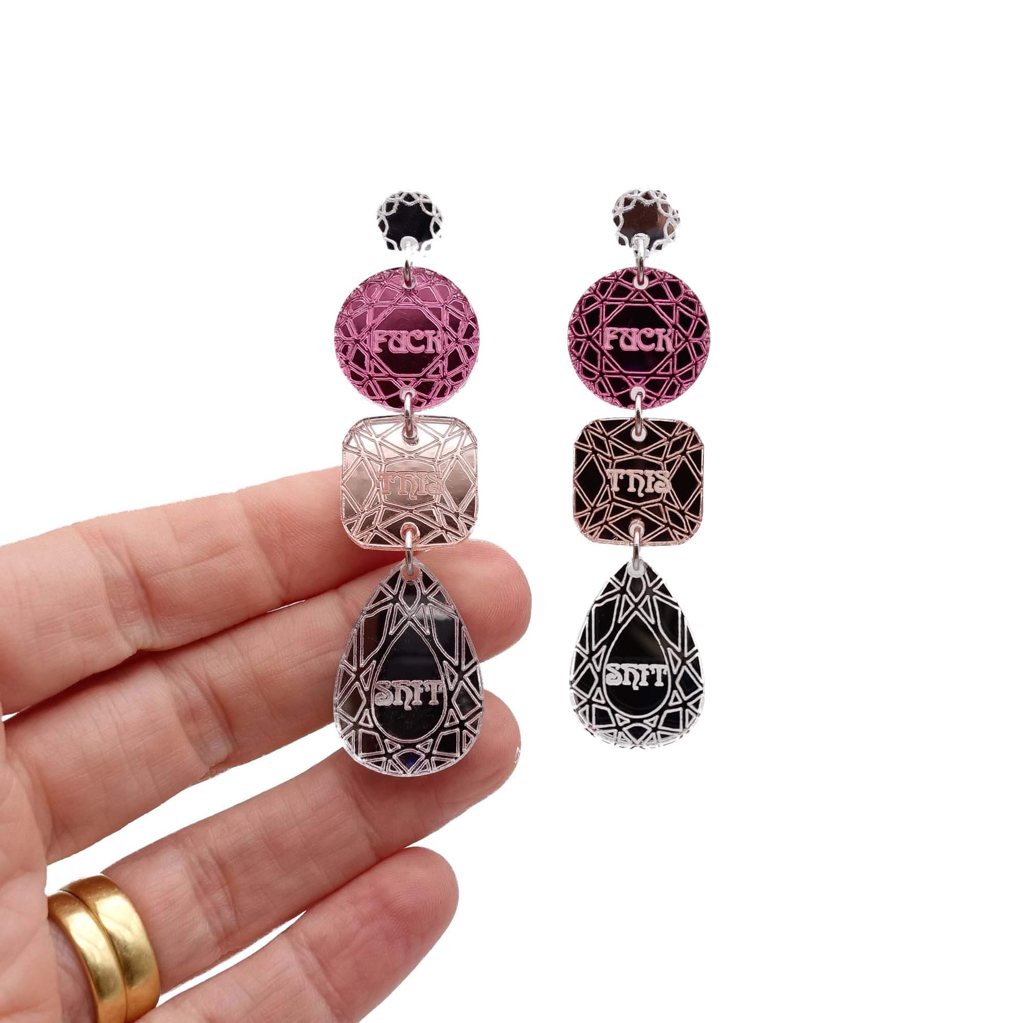 Sweary jewel drop earrings in pink, rose gold and silver, shown hanging against a white background with my hand behind.
