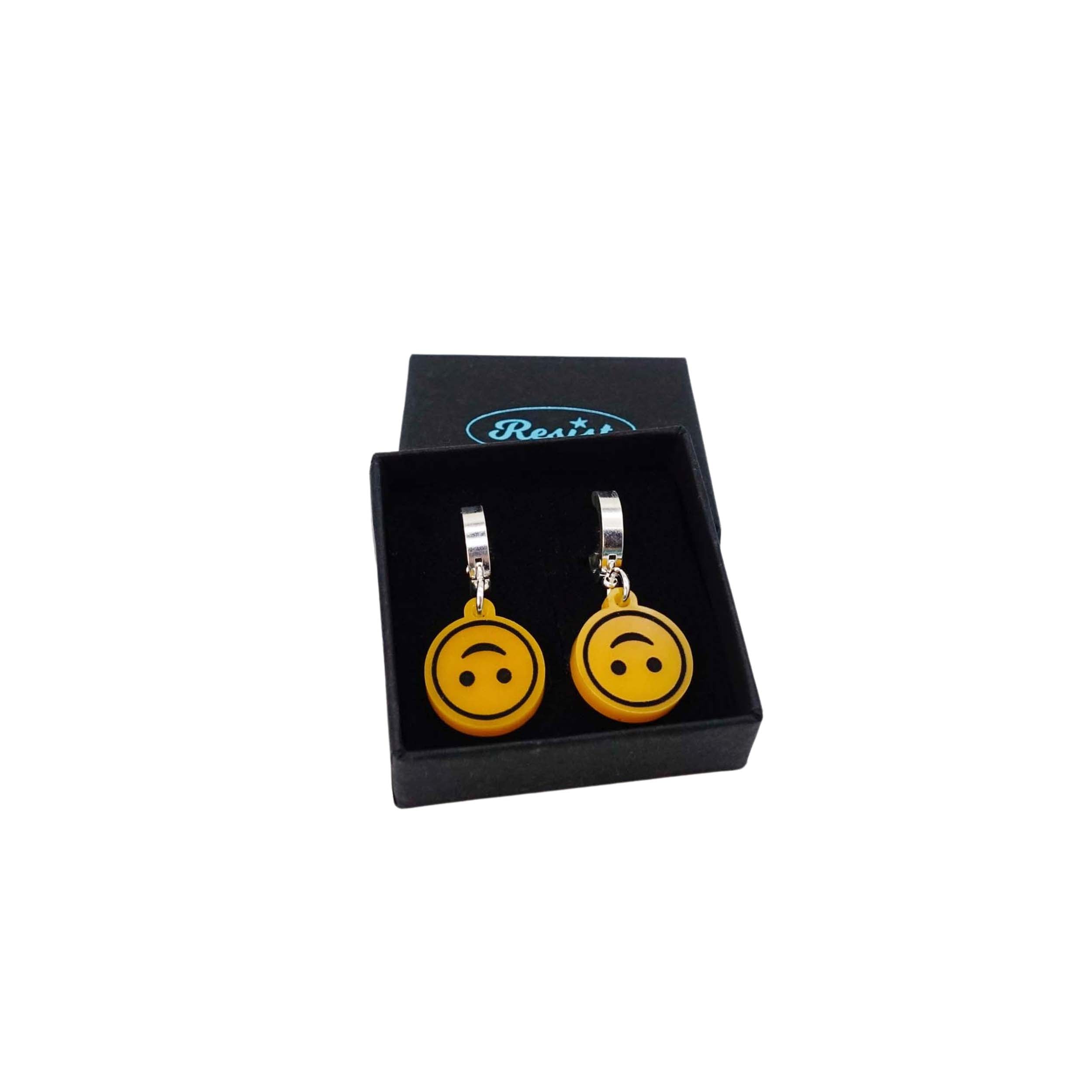 Upside-down smiley face emoji earrings on silver huggie hoops shown in a Wear and Resist gift box.