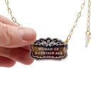 A hand holding a necklace with a tag that reads 'WOMAN OF A CERTAIN AGE' on a chain, finished with an iridescent lightning bolt charm.
