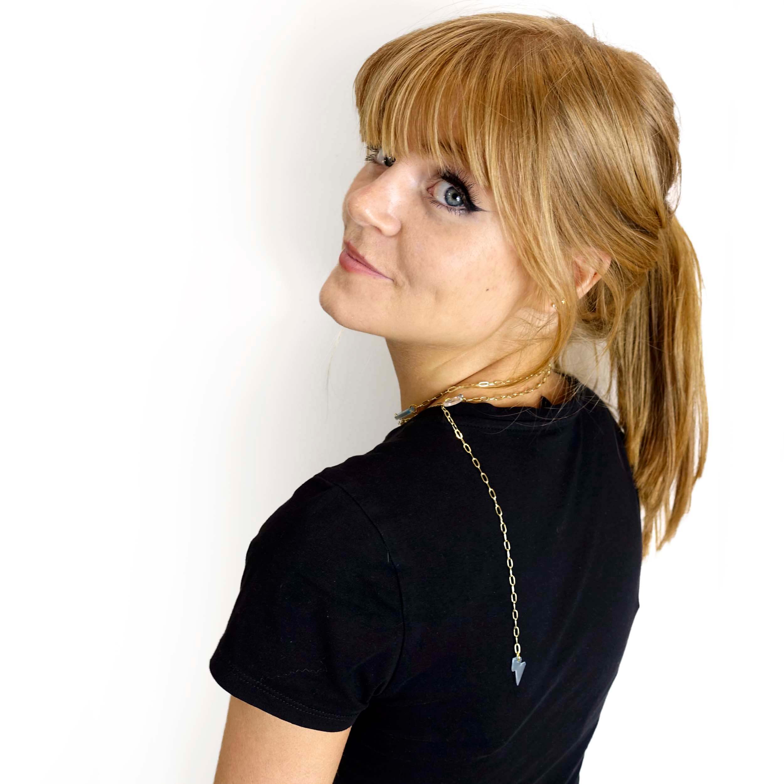 Eliza wears the Have a Word With Yourself design as a shorter necklace on a gold-plated paperclip chain, with the words 'persist' and 'be brave' etched in glow-in-the-dark pigment. She is turning to look over her shoulder, with the lightning bolt charm hanging down over her back.