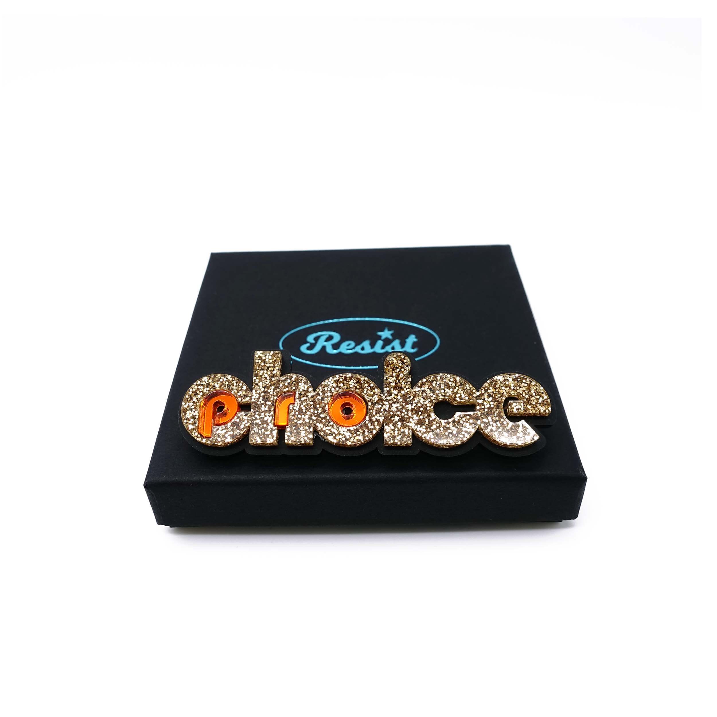Gold glitter and flame Pro-choice brooch, designed by Sarah Day for Wear and Resist, shown on a gift box.