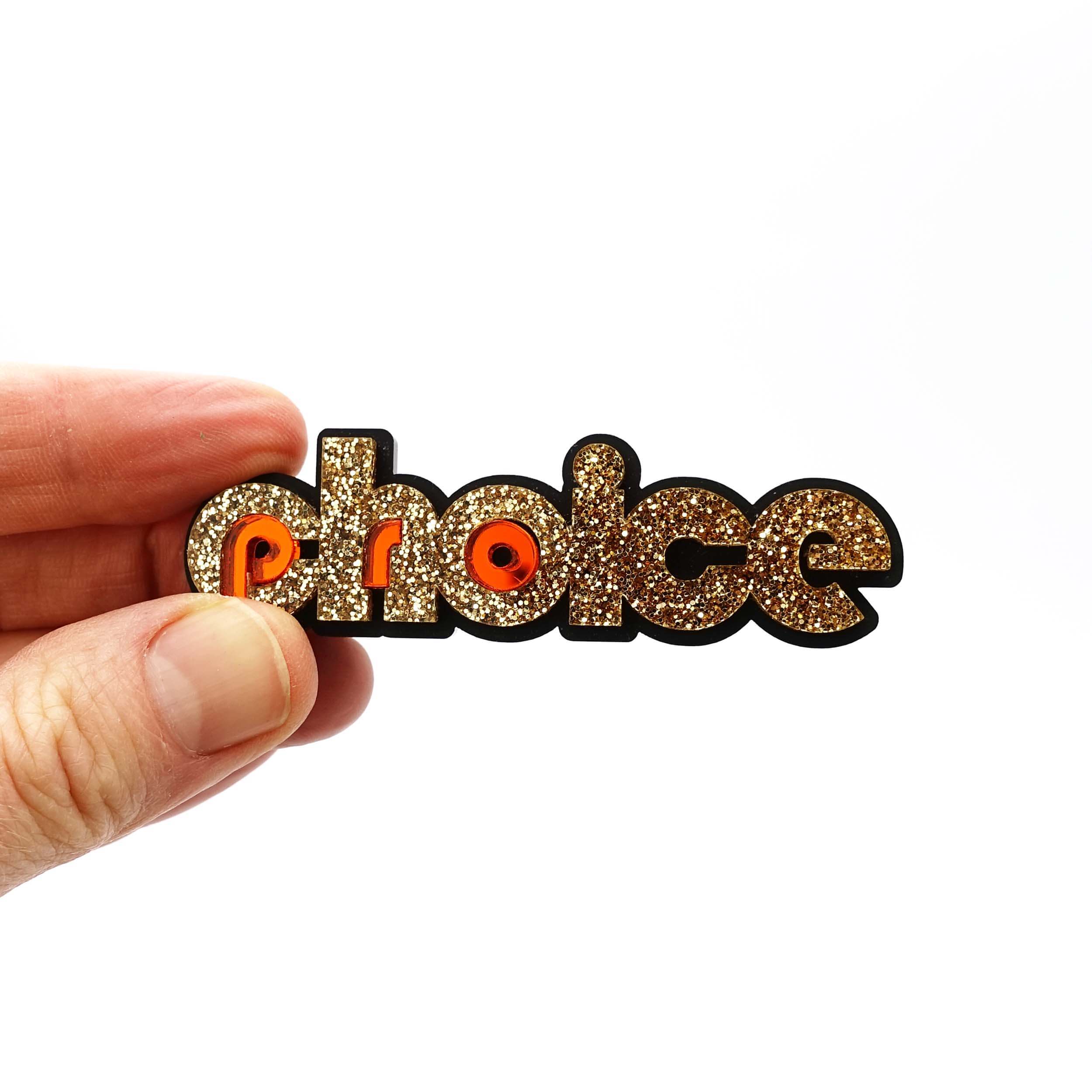 Gold glitter and flame Pro-choice brooch, designed by Sarah Day for Wear and Resist, shown being held in hand for size.