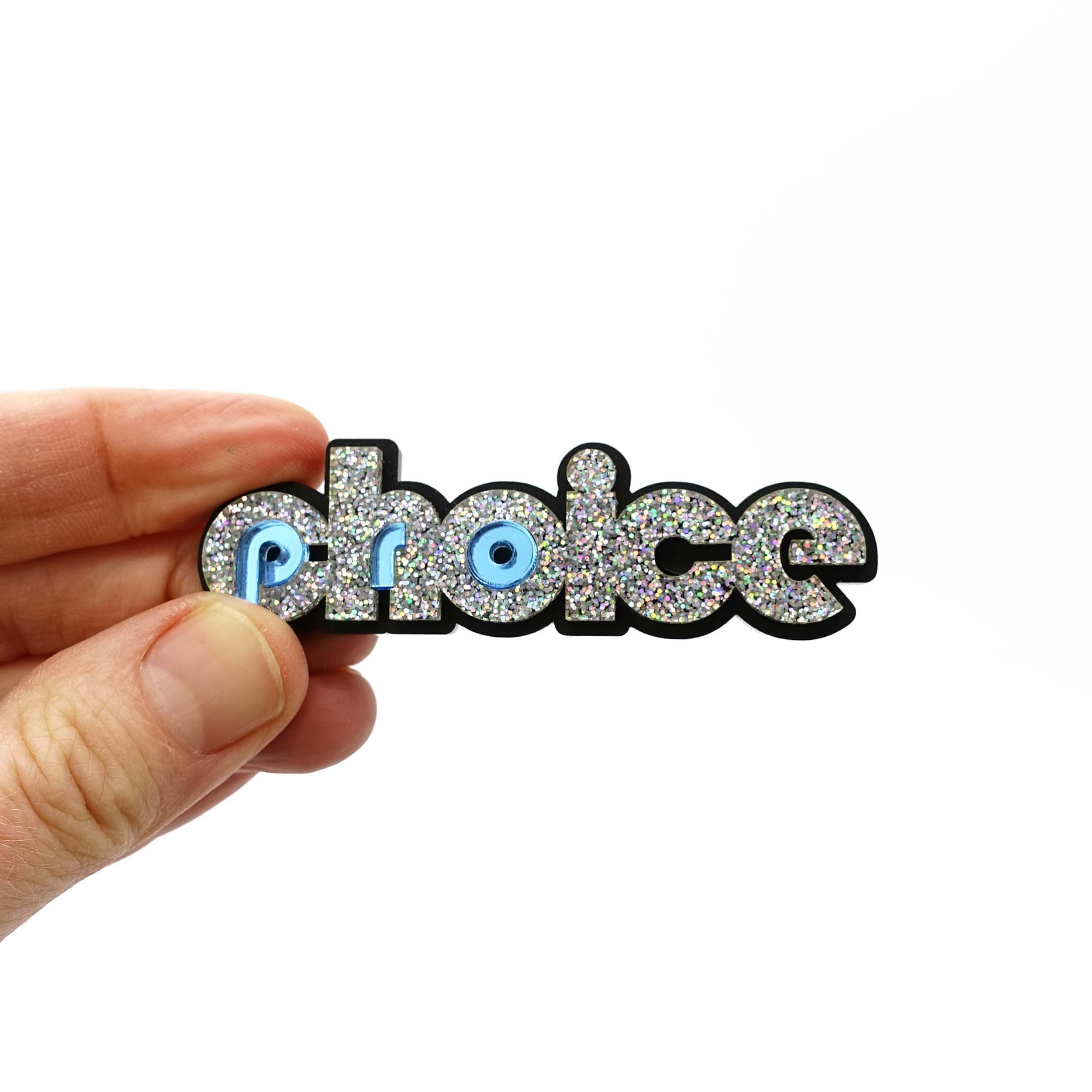 Iridescent silver glitter and sky mirror Pro-choice brooch, designed by Sarah Day for Wear and Resist, shown being held in a hand for size.