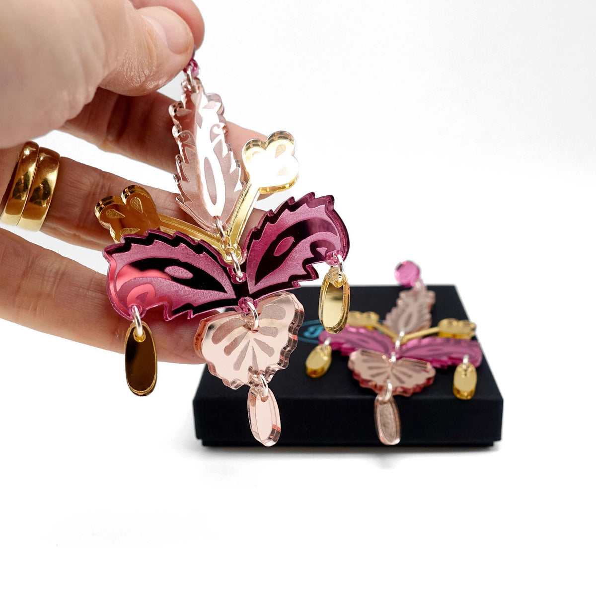 Rosey gold Festive Drop statement earrings shown being picked up from a Wear and Resist gift box. 