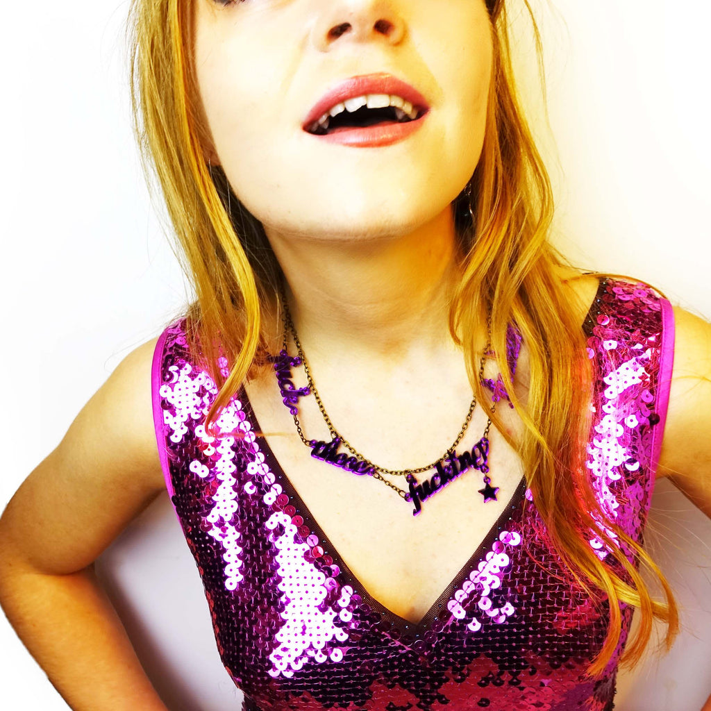 Model wears purple mirror f*ck these F*cking F*ckers necklace and a pink sequin dress. Designed by Sarah Day for Wear and Resist. 