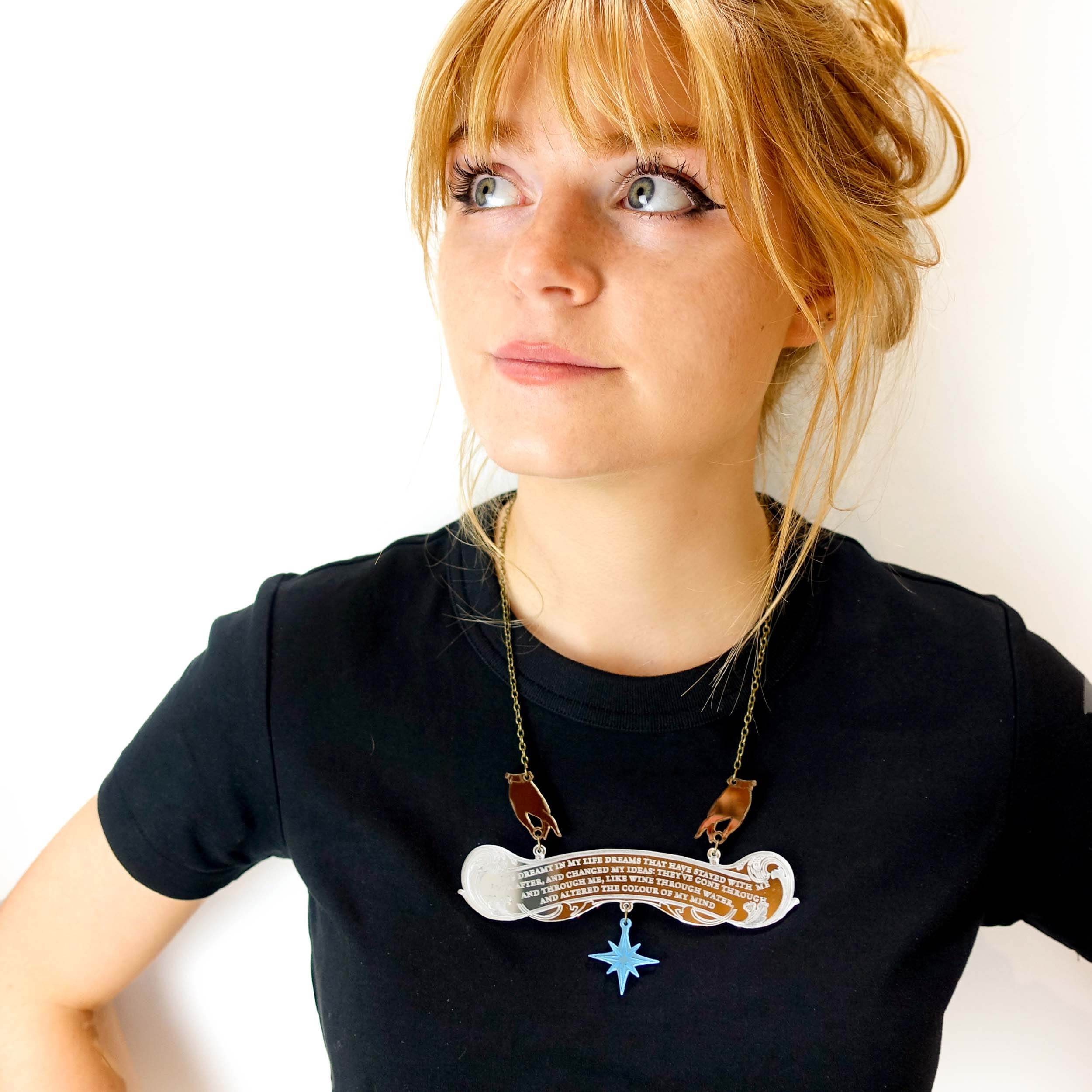 Eliza wears the Wuthering Heights Dreams necklace with a quote from Cathy etched on the silver mirror, held by two hands, and finished with a blue star. 