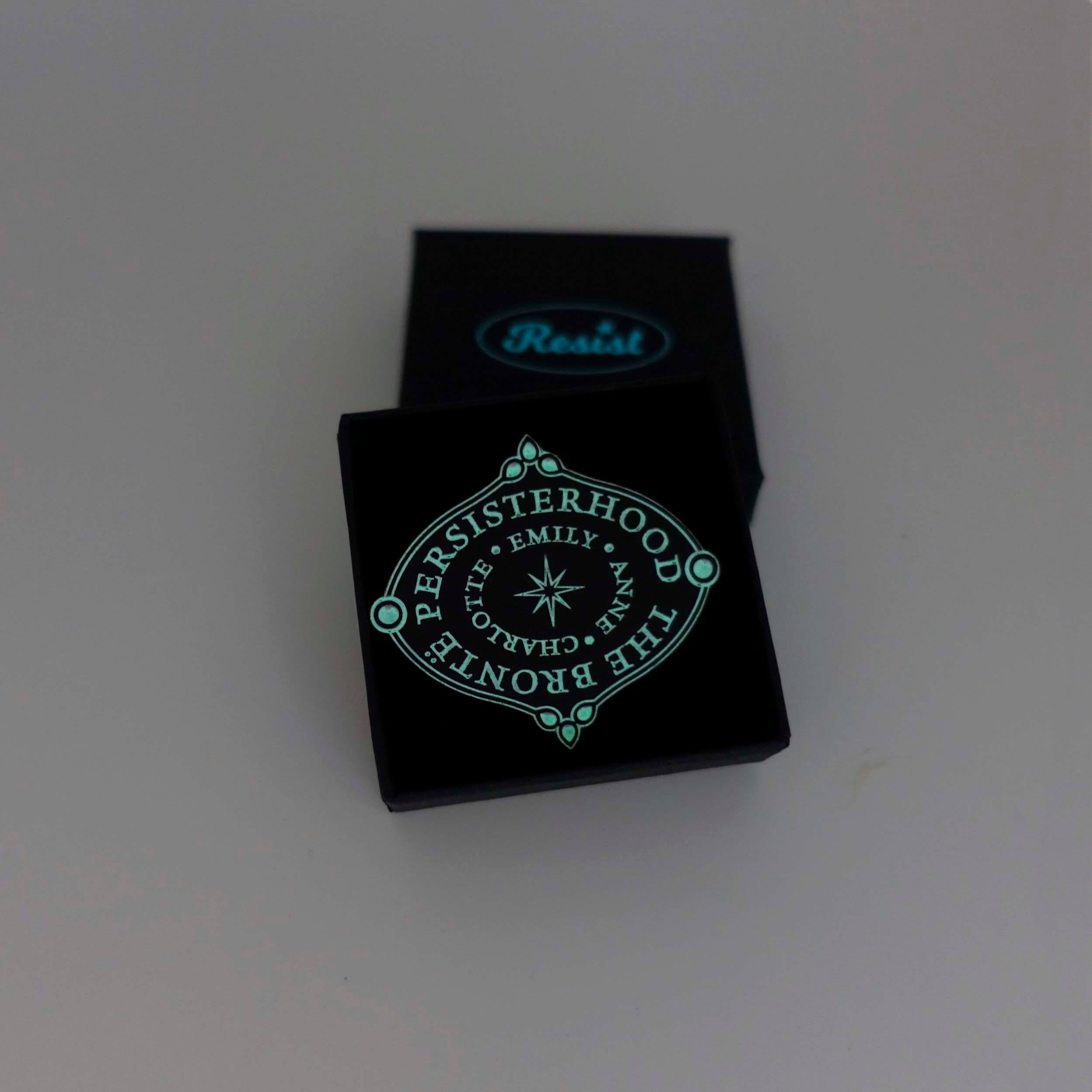 The Brontë Persisterhood brooch shown glowing in the dark in a Wear and Resist gift box.