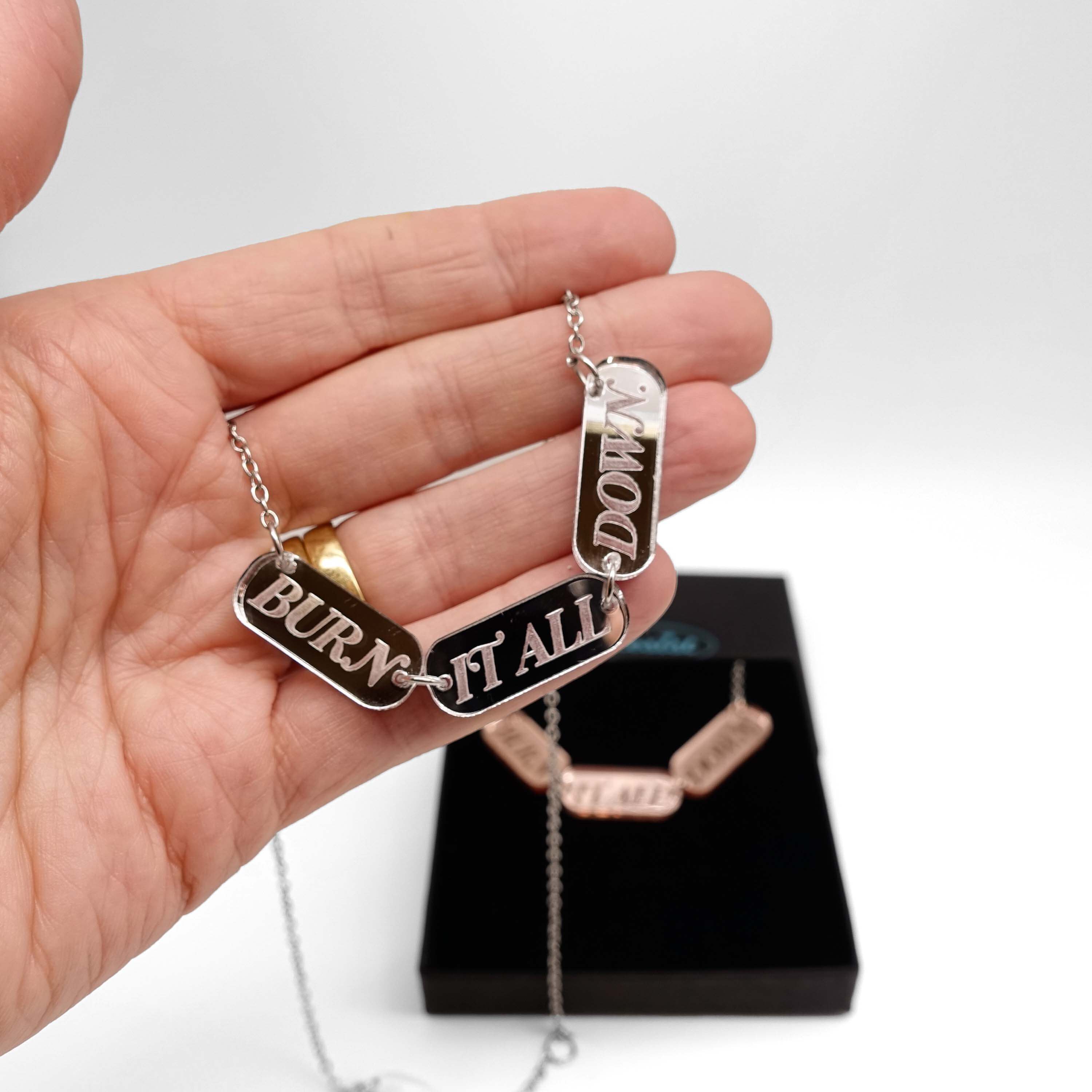 My hand holding a BURN IT ALL DOWN necklace in silver mirror on a stainless steel chain, with a rose gold version shown in the background in a Wear and Resist gift box. 