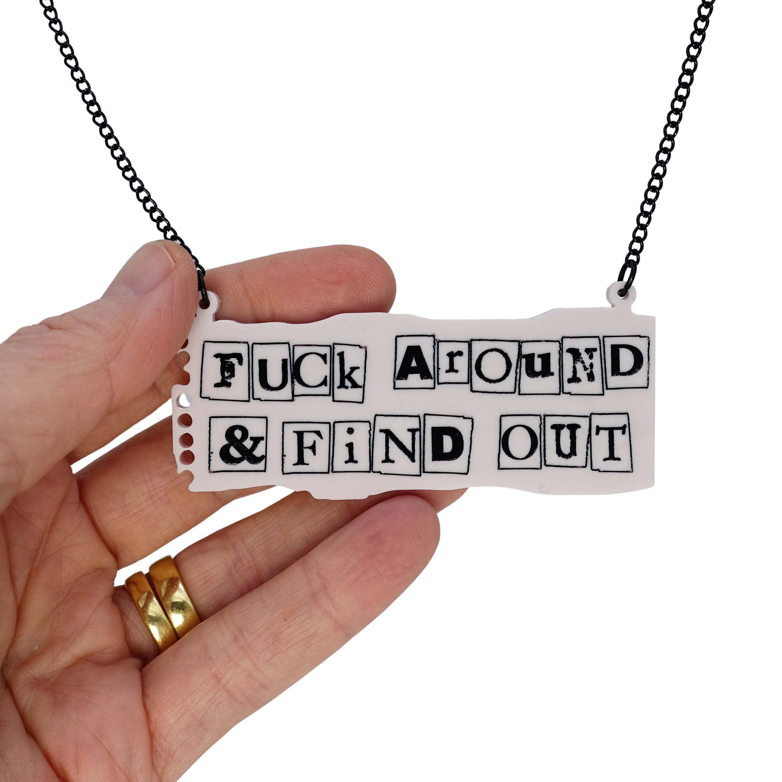 F*ck Around and Find Out necklace in the style of a ransom note on pale pink notepaper, shown hanging with my hand behind for scale. 