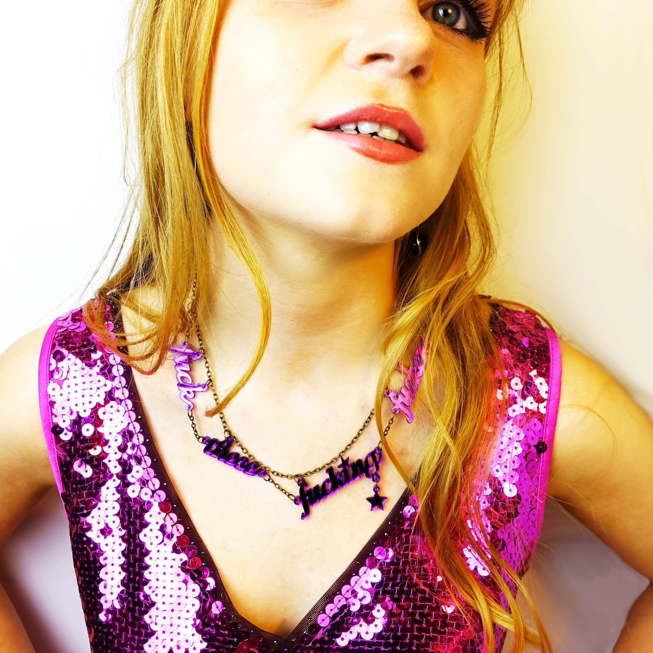 Model wears a sparkly pink dress and a poison lies purple fuck these fucking fuckers necklace designed and handmade by Sarah Day for Wear and Resist. £2 from all sweary jewellery goes to support Bloody Good Period. 