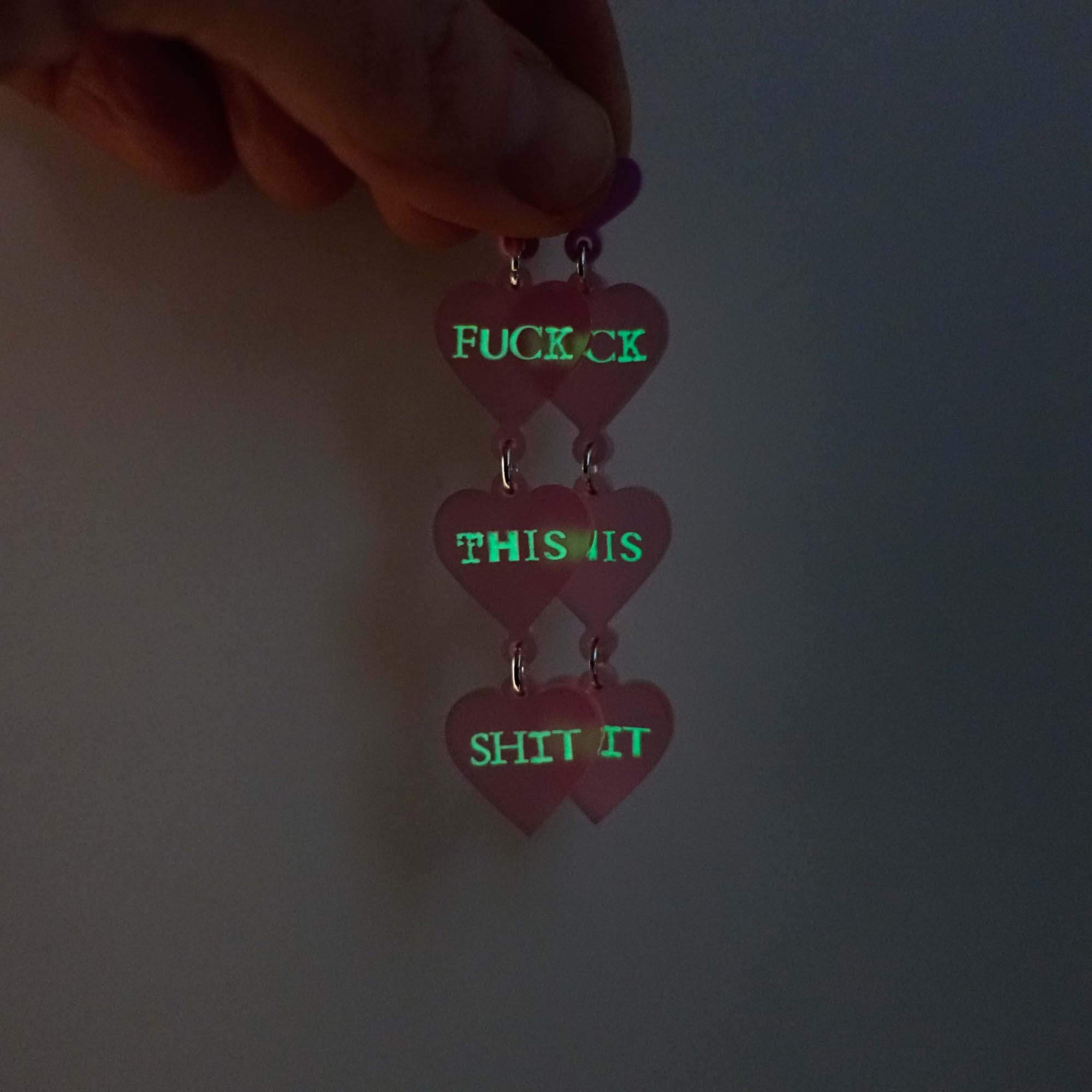 F*ck this Sh*t earrings in pale pink shown held up glowing in the dark. 