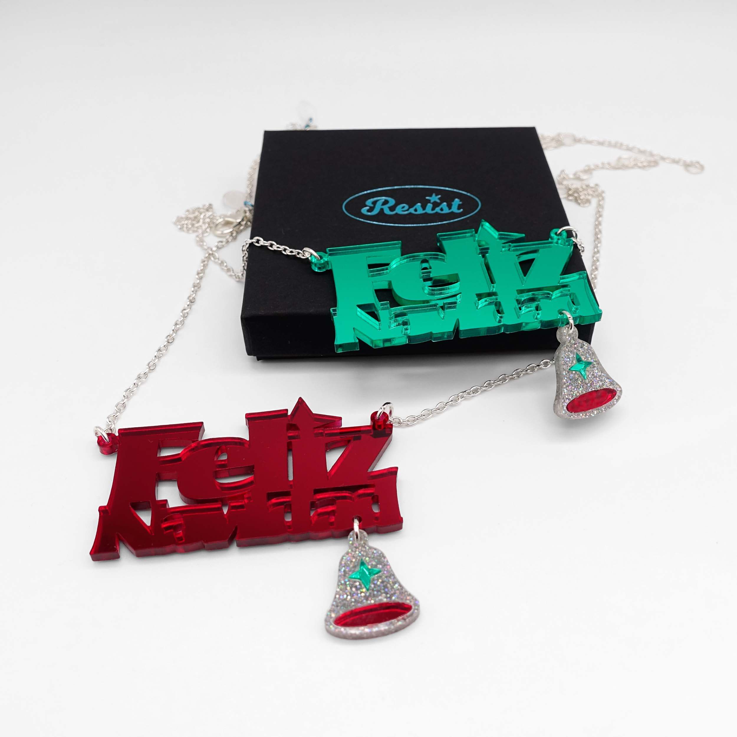 Both colours of the Feliz Navidad necklace shown together: electric green and ruby red mirror, with one resting on a Wear and Resist gift box.