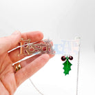 My hand holding the Festive AF matte iridescent necklace with hanging holly leaf and berries on a stainless steel chain. 