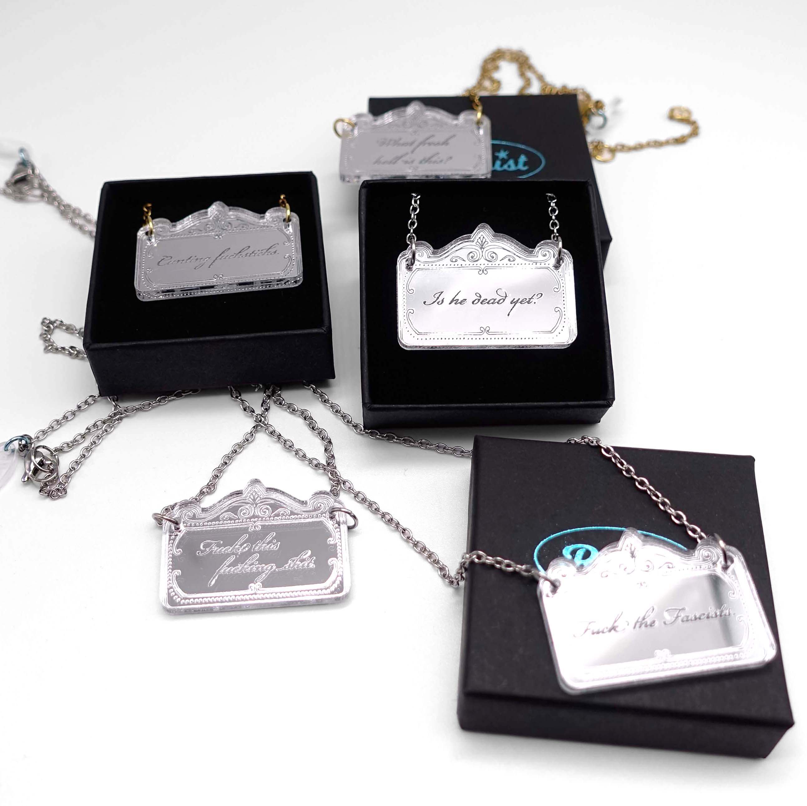 A group of delicate etched necklaces etched with rude and sweary words shown in and on Wear & Resist small gift boxes. 