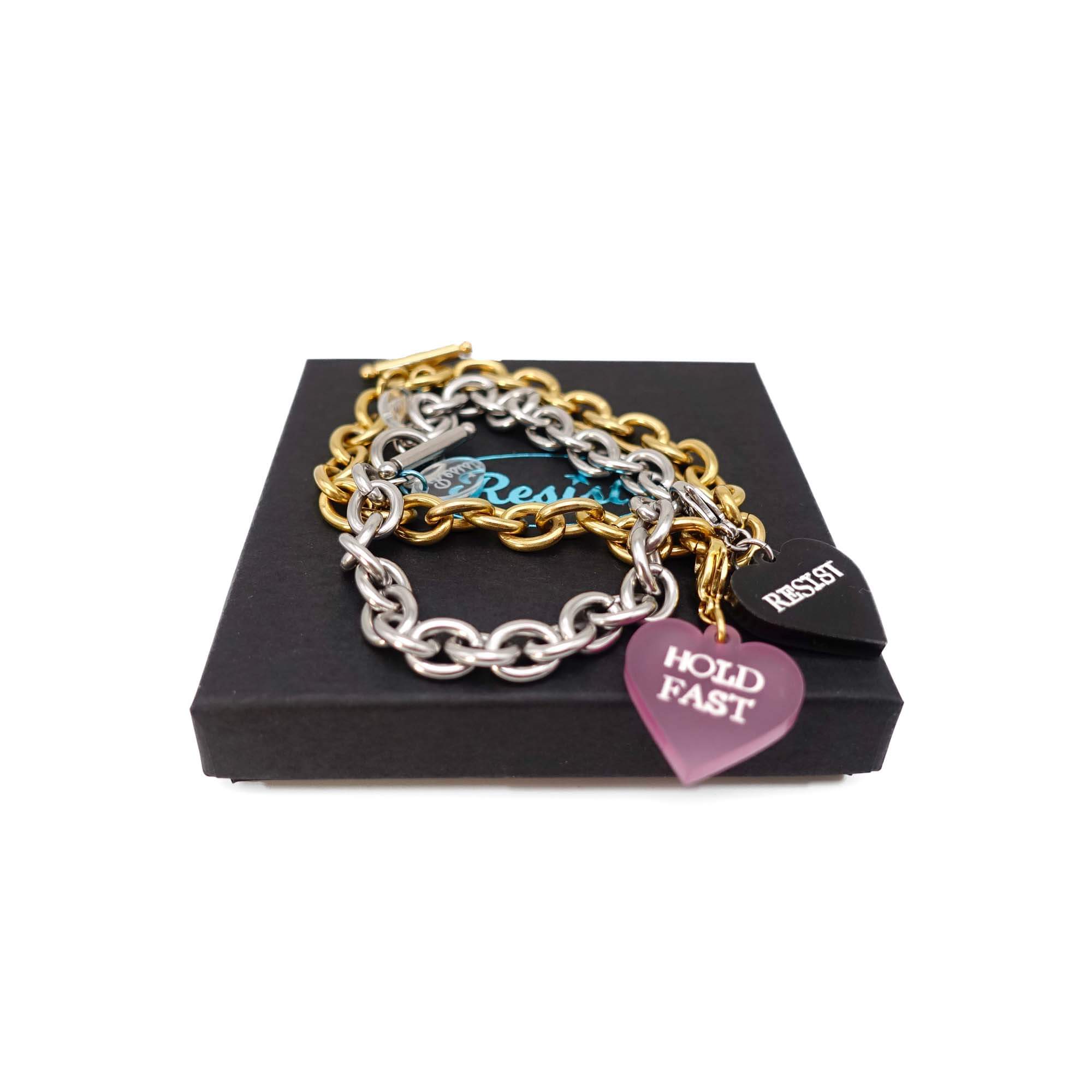 Charm bracelets in stainless steel and gold shown on a Wear and Resist gift box with Hold Fast and Resist charms that glow in the dark. 