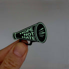 Megaphone brooch etched iwith the words Hope Not Hate, glowing in the dark. 