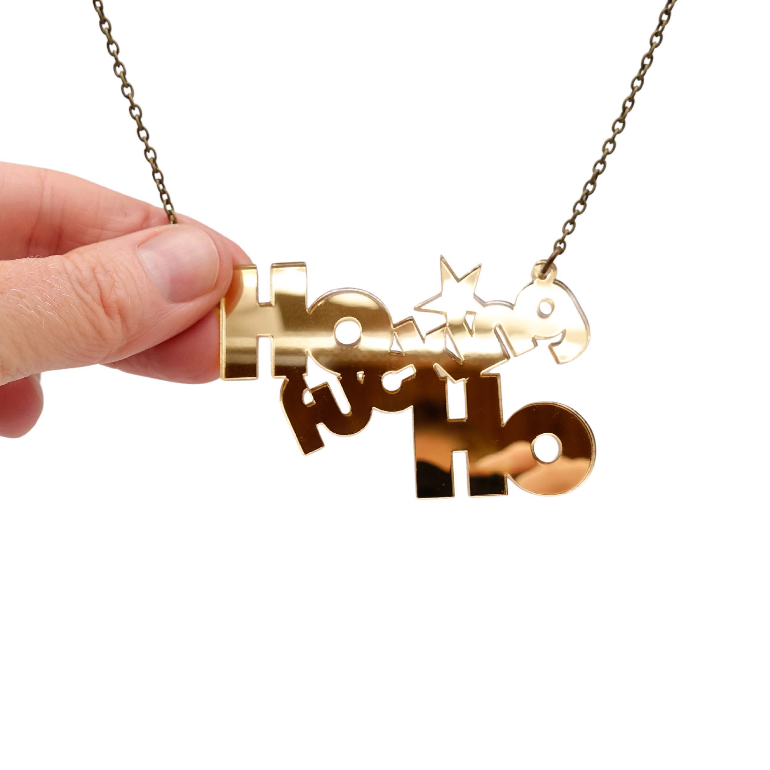 Ho F*cking Ho necklace in gold mirror, shown hanging against a white background with my hand holding it for size and to show the reflective quality of the mirror. 