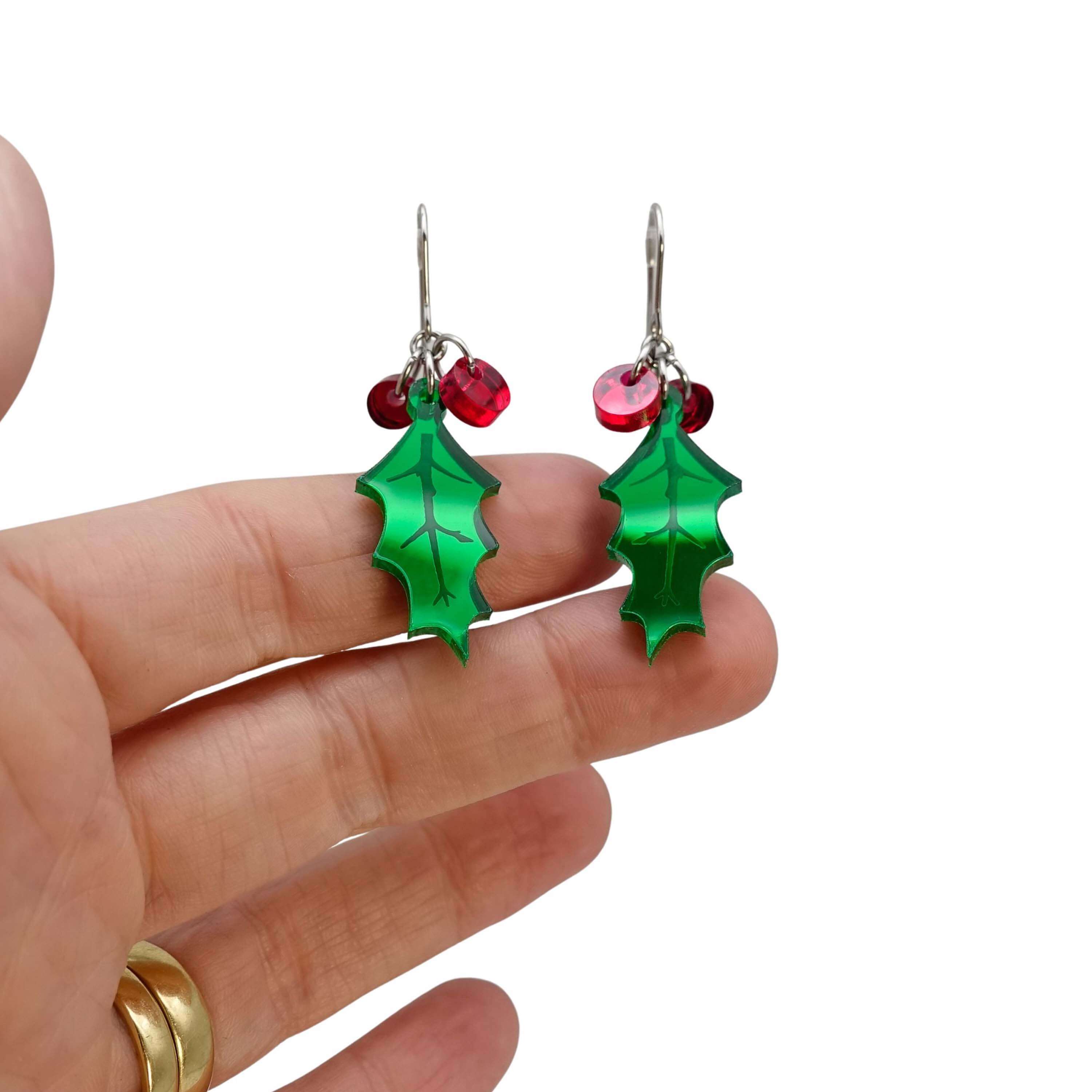 Naturalistic Holly earrings with red berries shown hanging on stainless steel ear wires against a white background with my hand behind for scale. 
